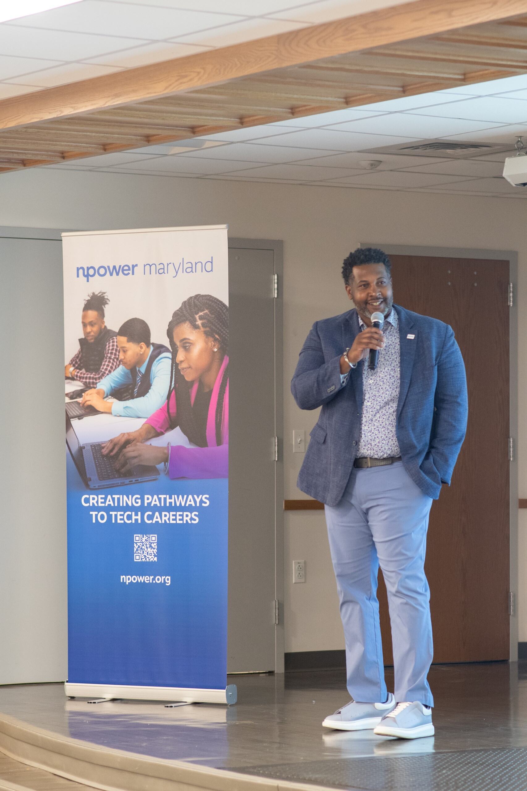 A man speaks into a microphone on stage beside a banner that reads "NPower Maryland: Creating Pathways to Tech Careers" with images of people using laptops.