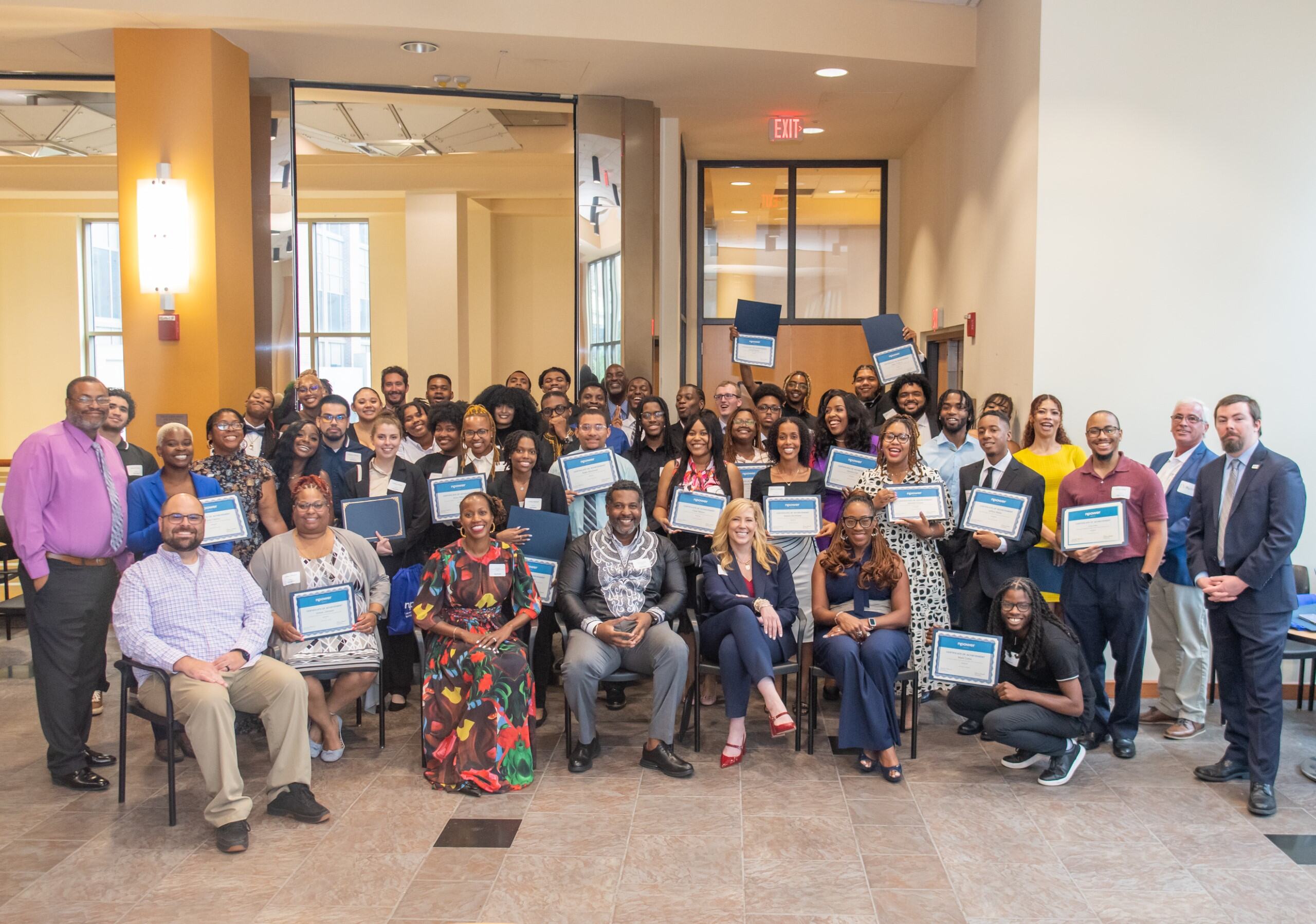 A large group of people, some seated and some standing, gather indoors holding certificates. They're smiling at the camera.