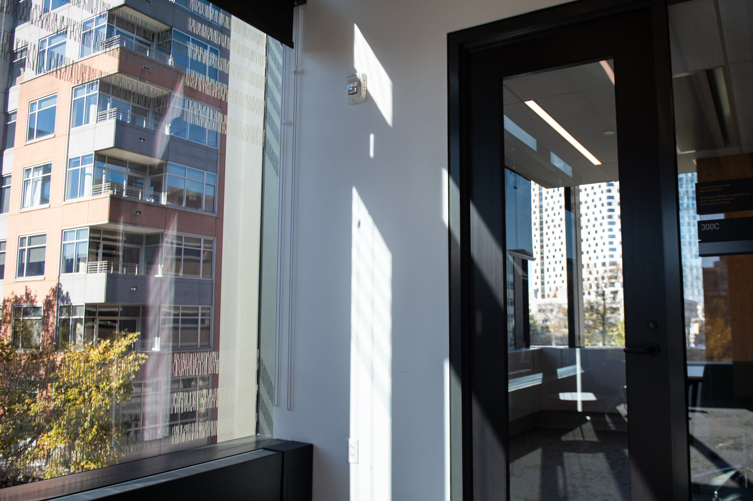 Sunlight streams through a large window in an office space, illuminating a wall. A black interior door is slightly open, and an apartment building is visible outside.