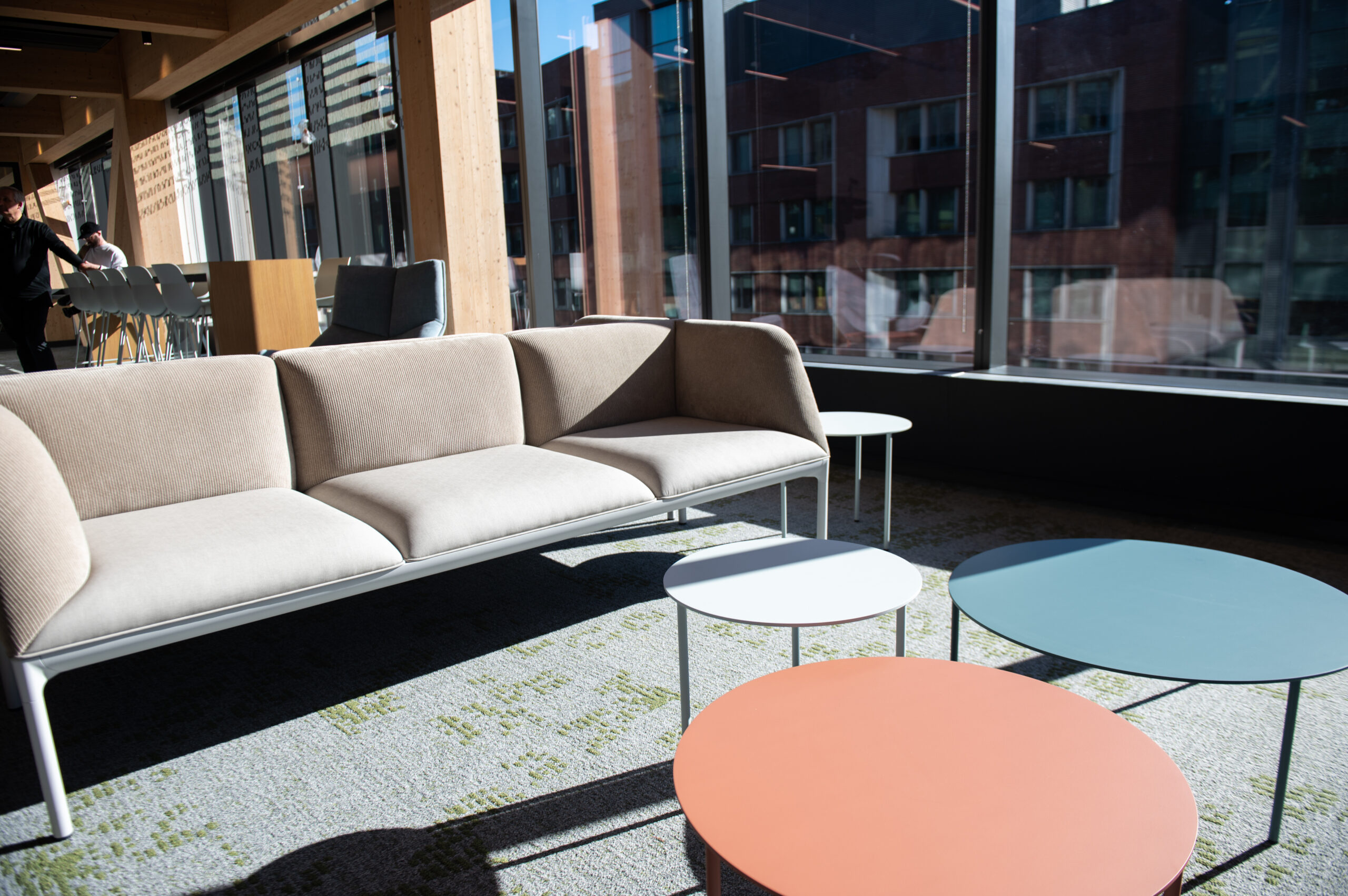 A modern office lounge with a beige sofa and round, colorful coffee tables in front of large windows. Building exterior visible through the windows.