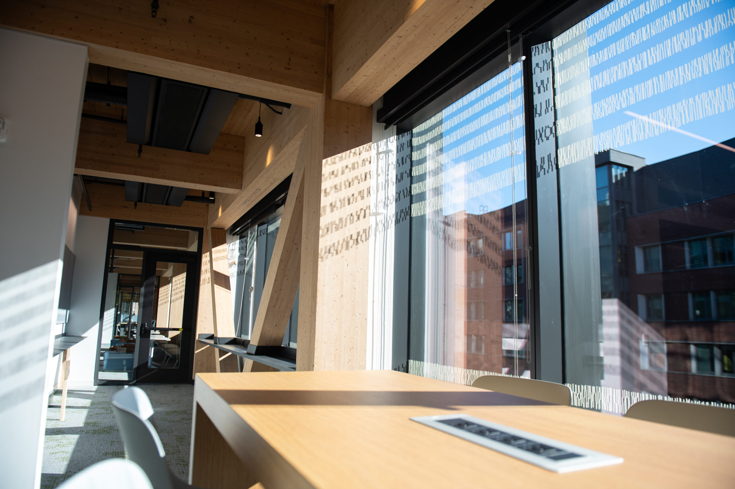 Sunlit modern office space with large windows, wooden furnishings, and a table with chairs. Building exteriors visible outside.