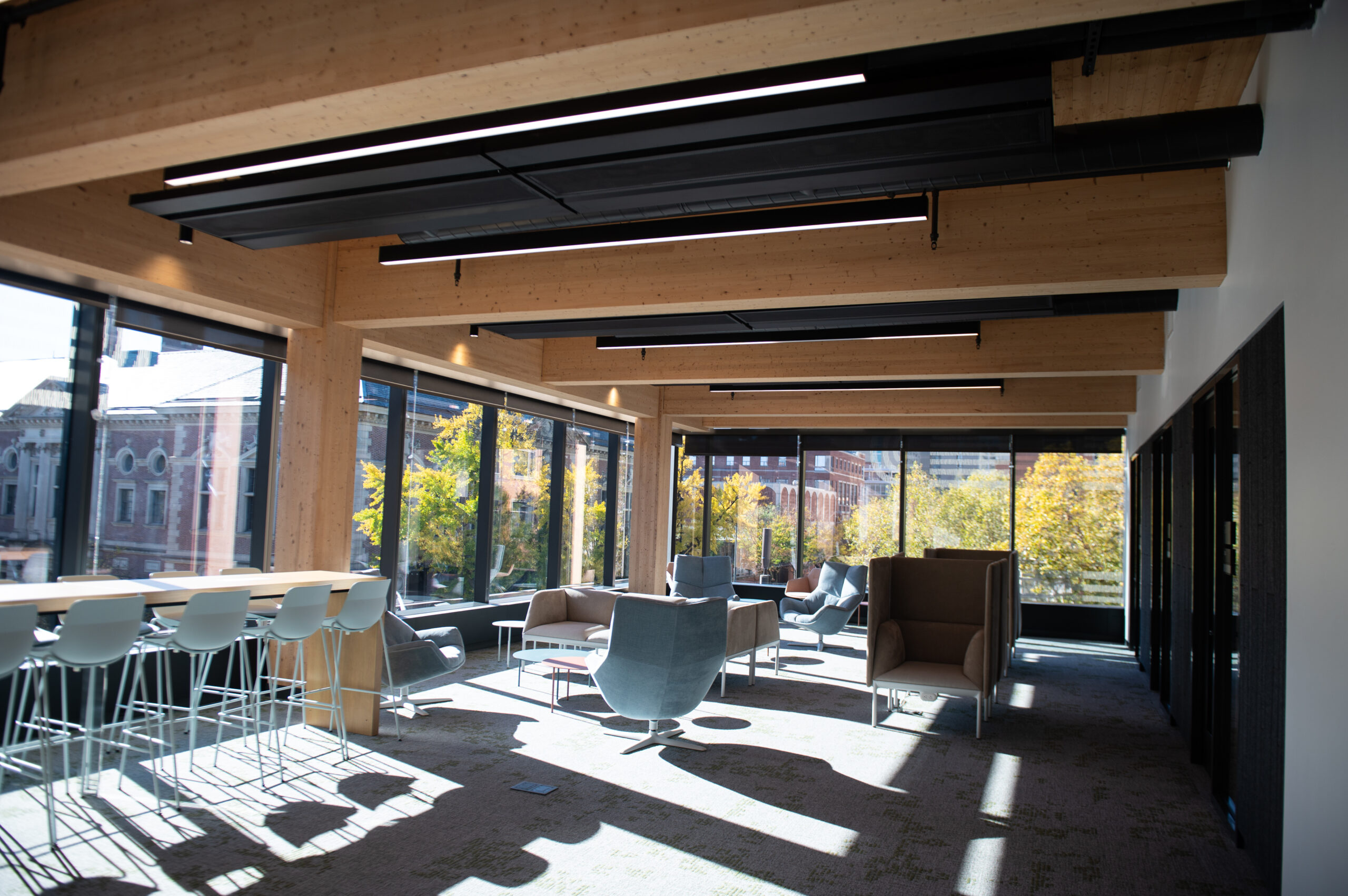 Modern office lounge with large windows, wooden ceiling beams, and various seating options, including bar stools and armchairs. Trees and buildings are visible through the windows.