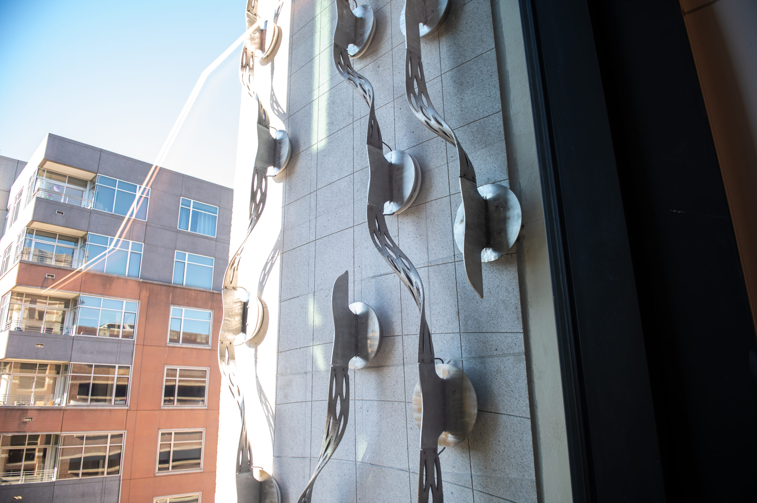 Close-up of a modern building facade with vertical metal wave-like sculptures and circular elements, seen from a window. A neighboring building is visible on the left.