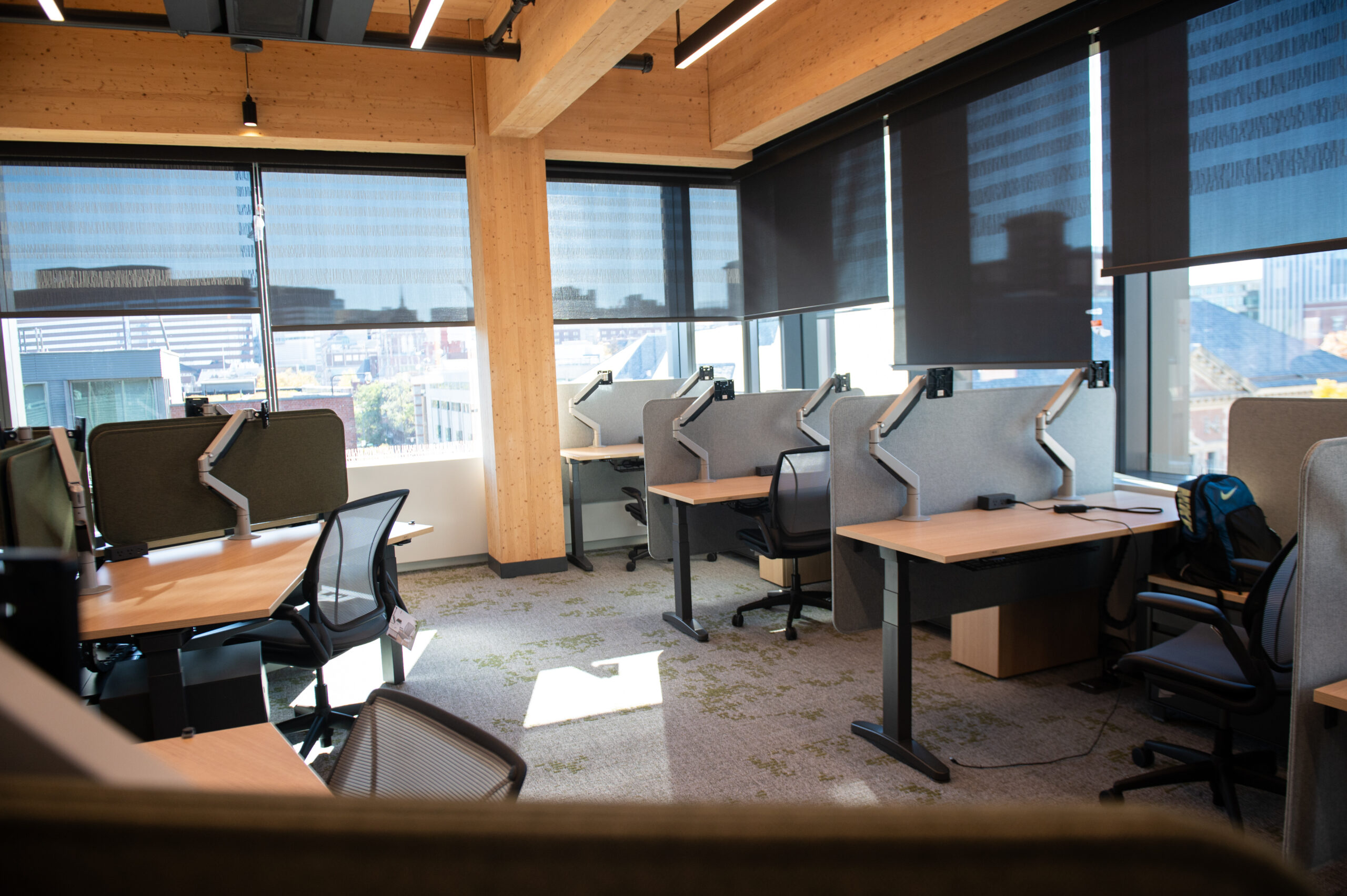 An empty office with several workstations, featuring modern desks, chairs, dividers, and large windows with city views and blinds.