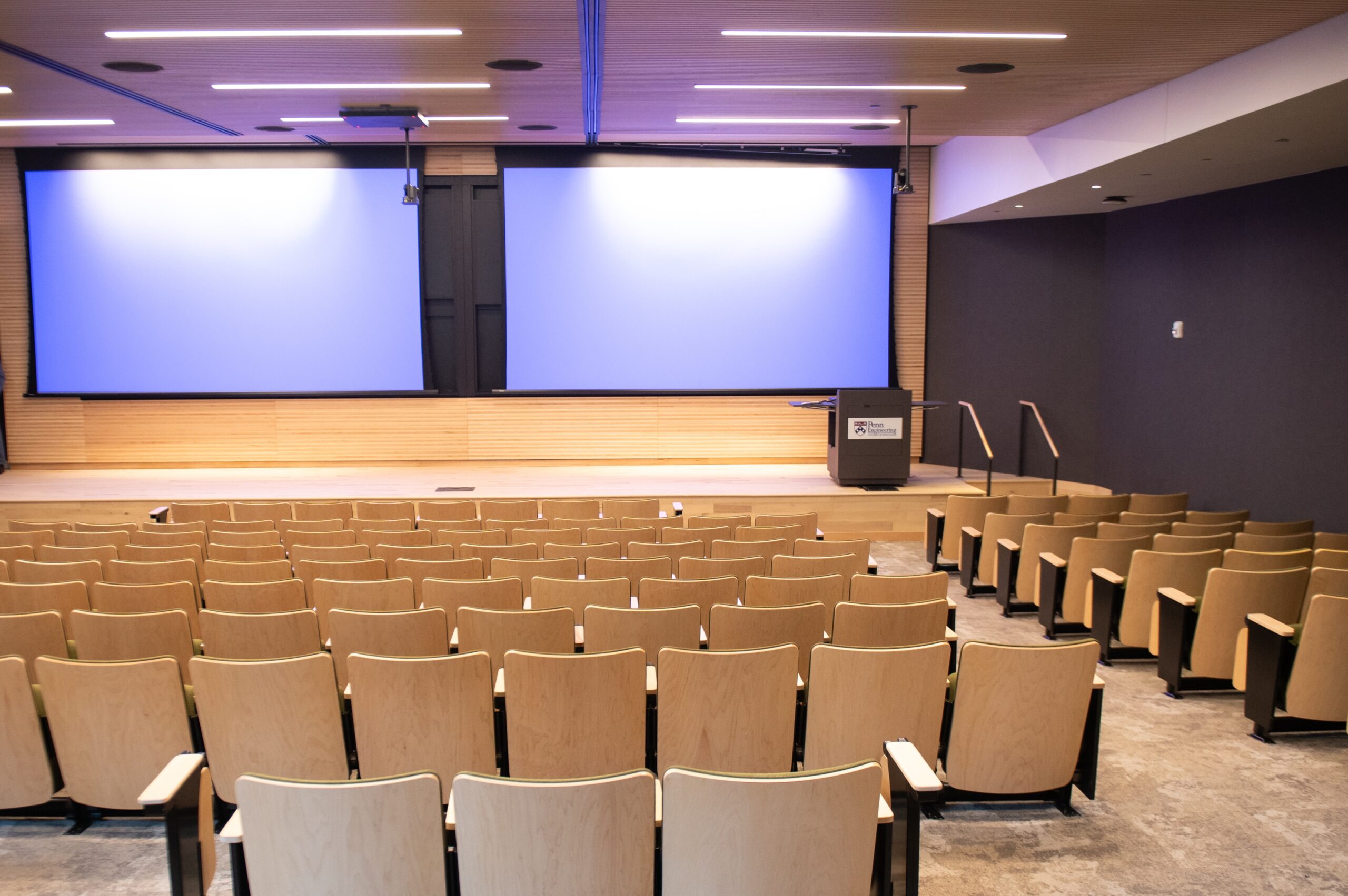 An empty lecture hall with rows of beige seats facing a stage with two blank projection screens.