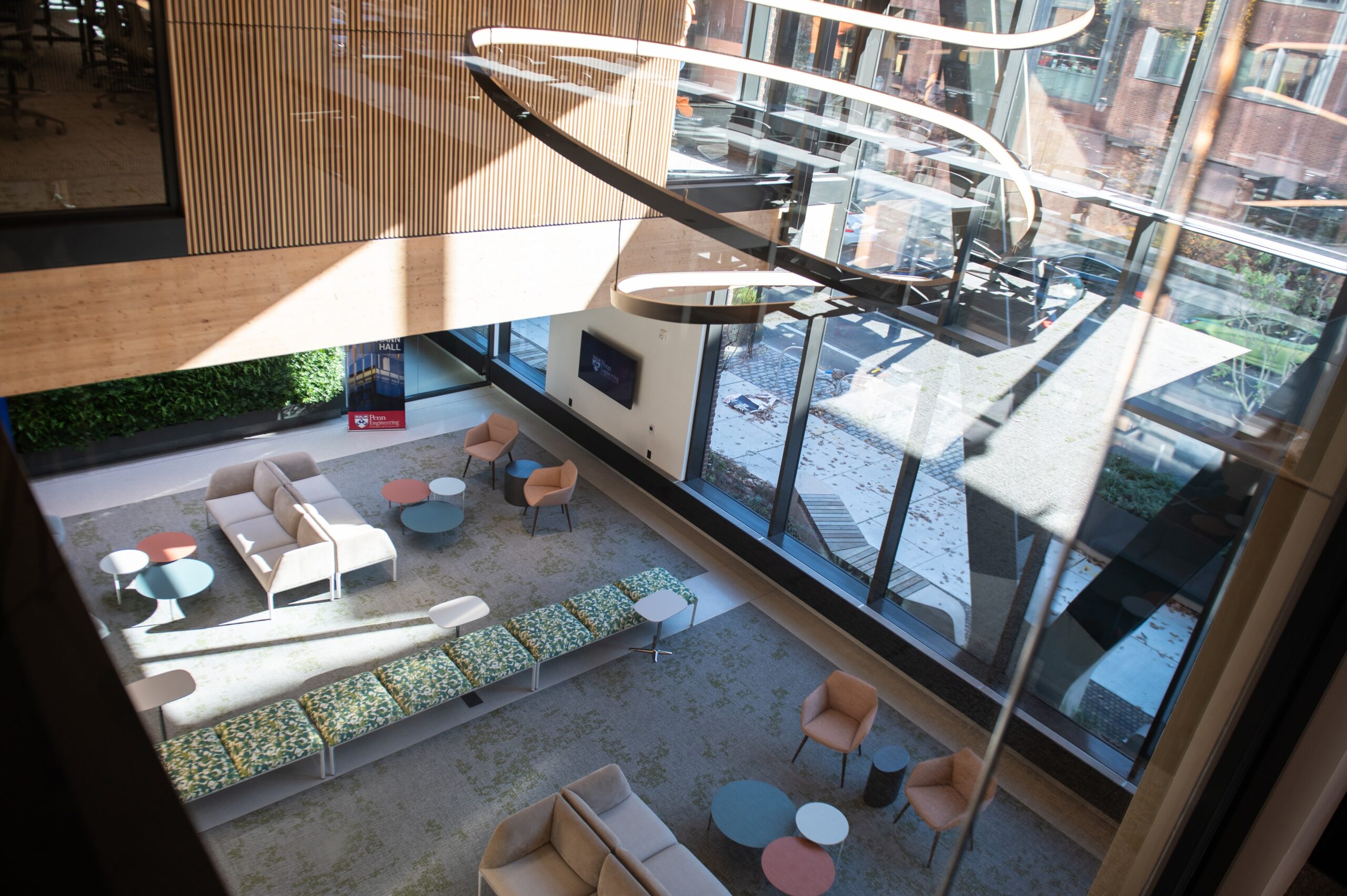 Modern atrium with large windows, couches, chairs, round tables, and artistic ceiling lights, overlooking a city street.