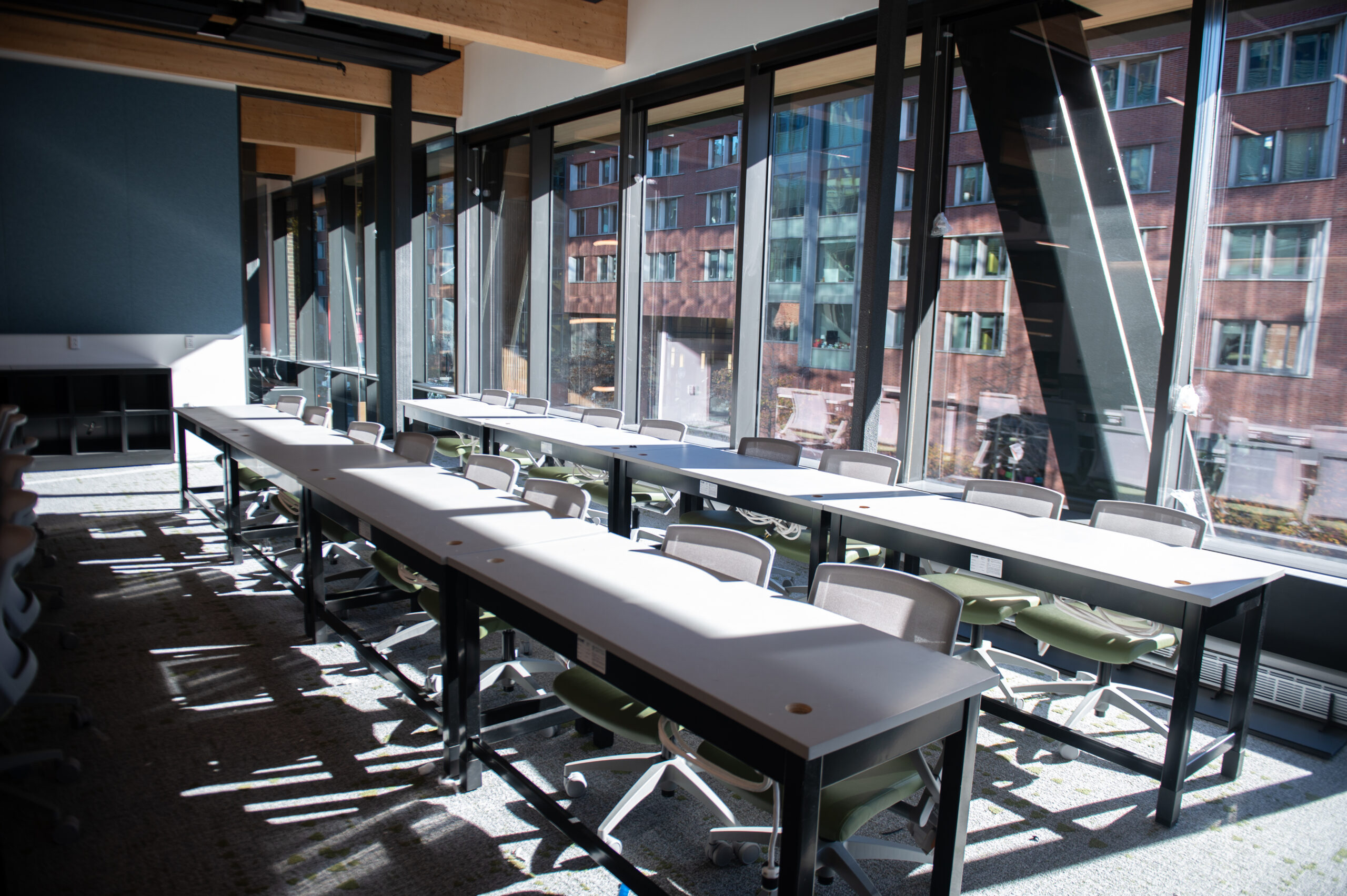 A modern classroom with long white tables and green chairs, set against a wall of large windows. Sunlight streams in, illuminating the room.