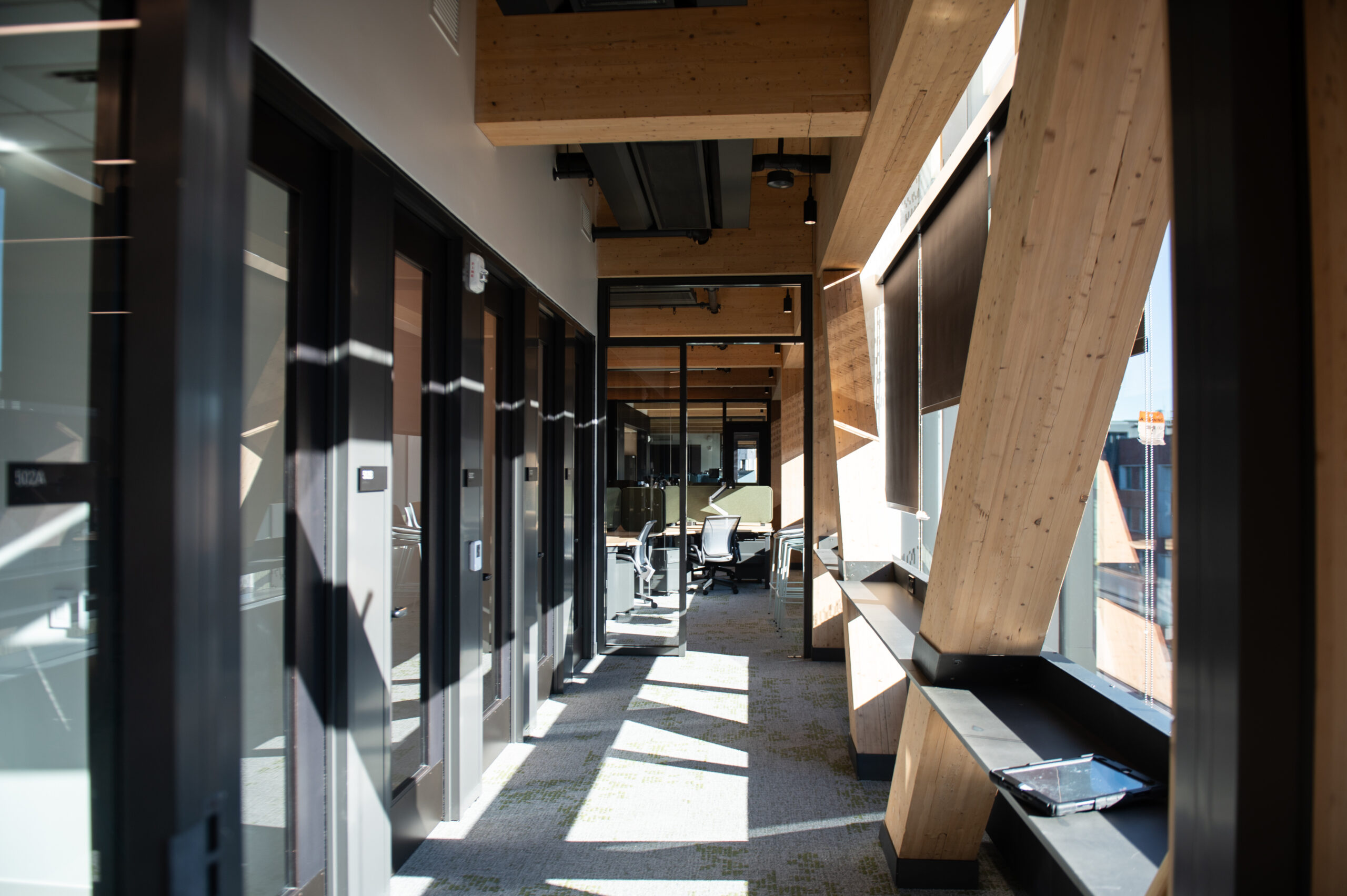 Sunlit office hallway with large wooden beams, glass-walled conference rooms, and a bench with a folder.