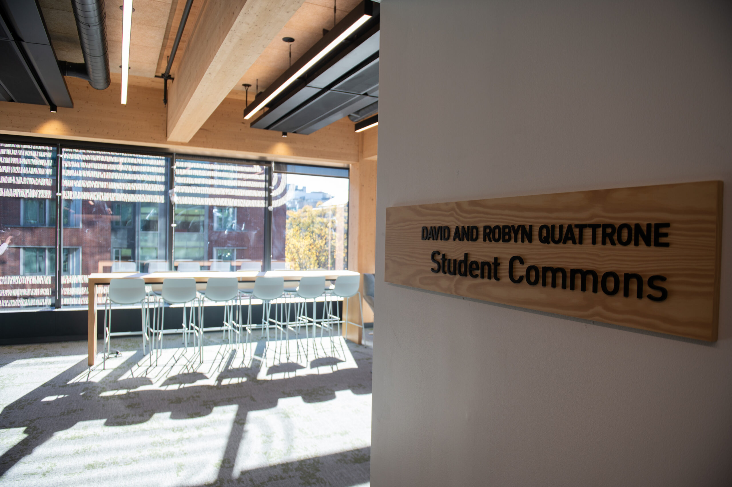 Modern student commons area with tall glass windows, bar-style table, and chairs. Sign reads "David and Robyn Quattrone Student Commons.