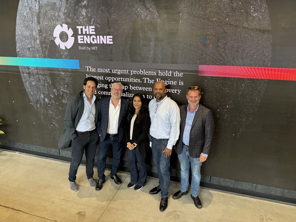 Five people standing in front of a wall with "The Engine Built by MIT" logo and text.