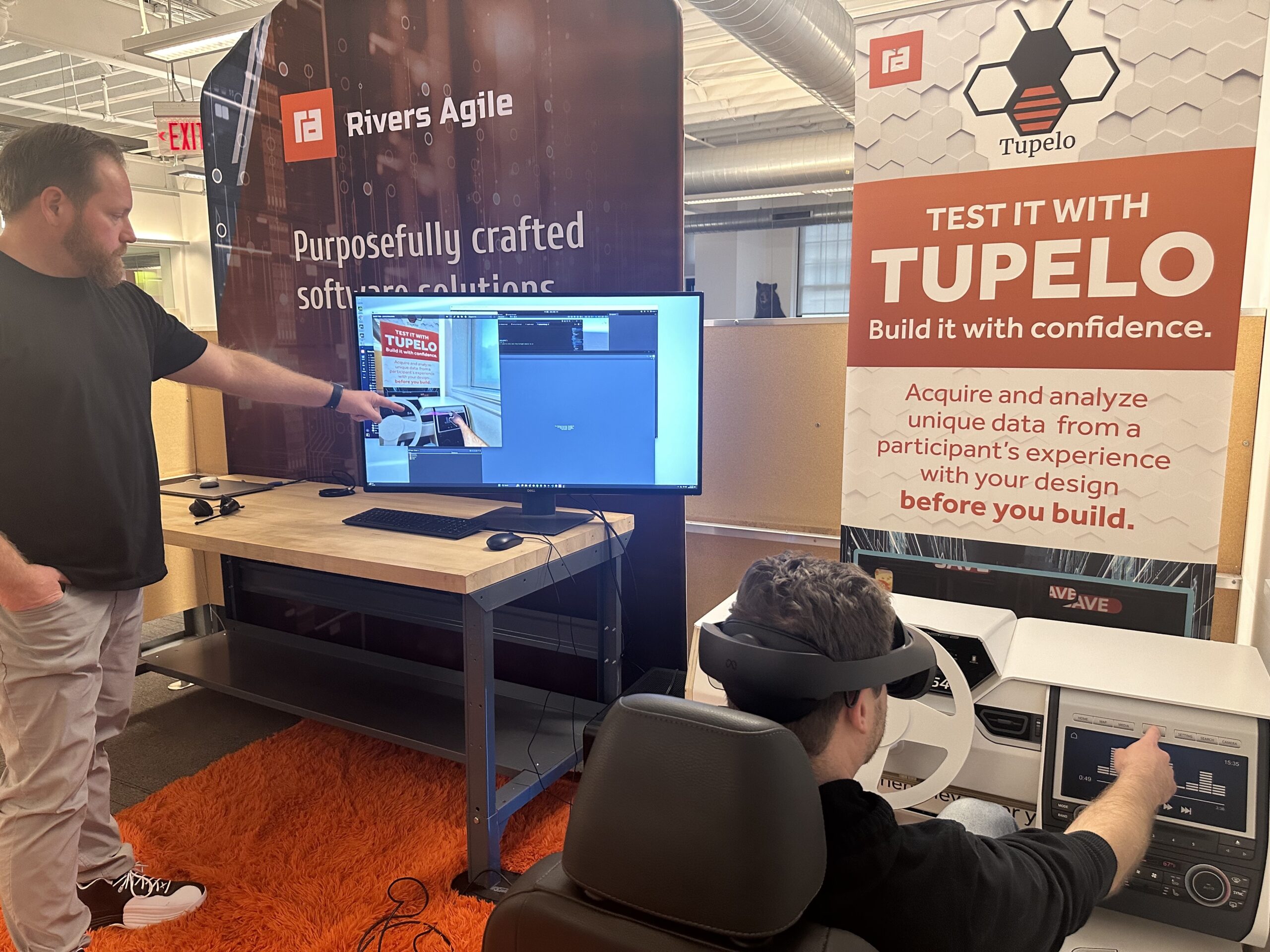 A man sits at a fake steering wheel wearing a VR headset, while another points at what he is seeing on a computer. 