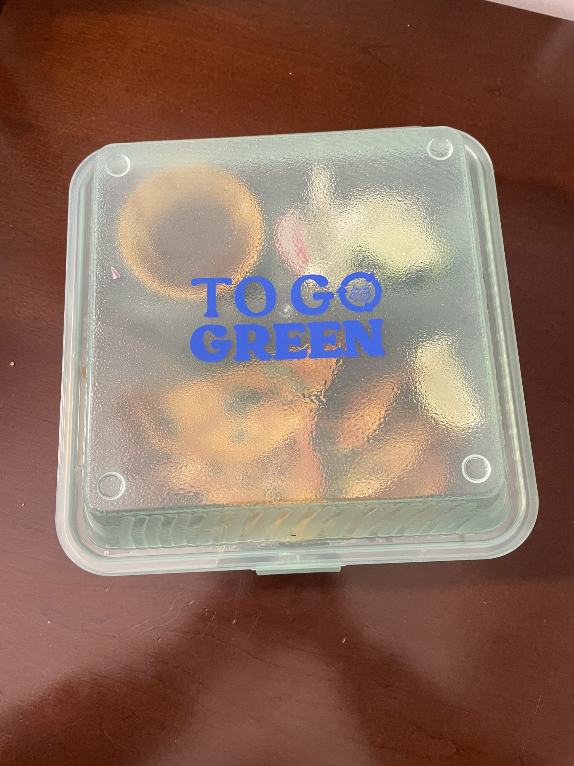 A frosted glass food container with pastries inside, labeled "To Go Green," sits on a wooden surface.