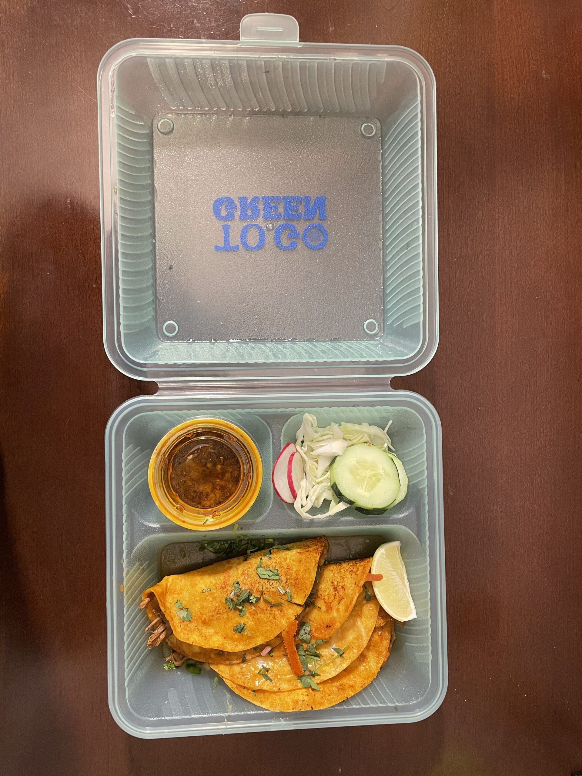 Takeout container with two tacos, a lime wedge, a slice each of cucumber and radish, and a small cup of sauce.