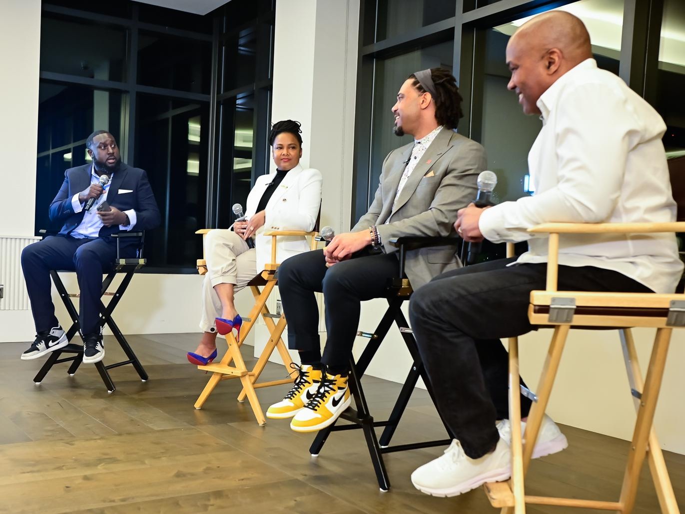 Three Black men and one Black woman sit at directors chairs in room with black windows and wood floor