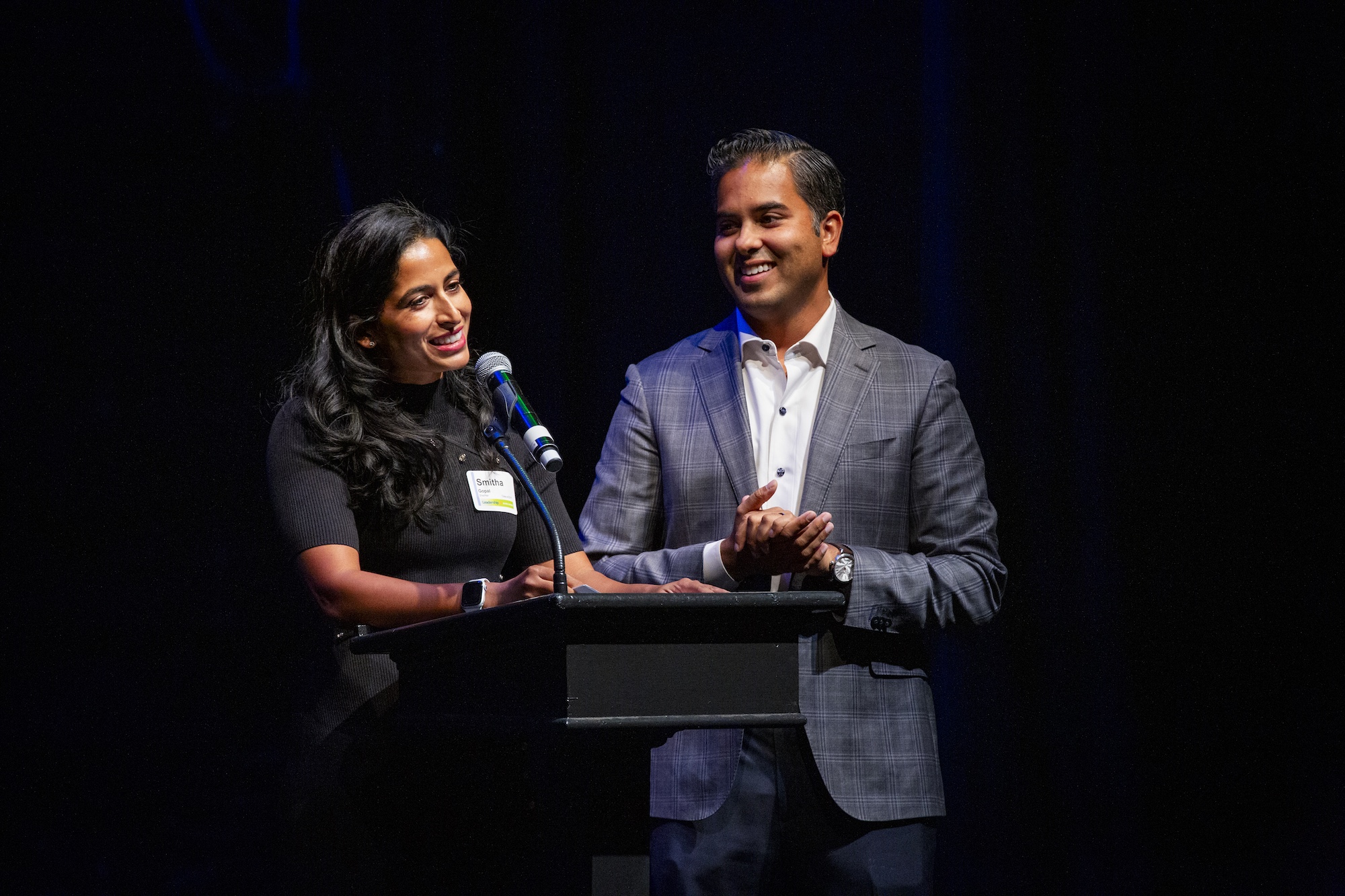 A woman in black anda. man in a grey suit stand before a black podium.