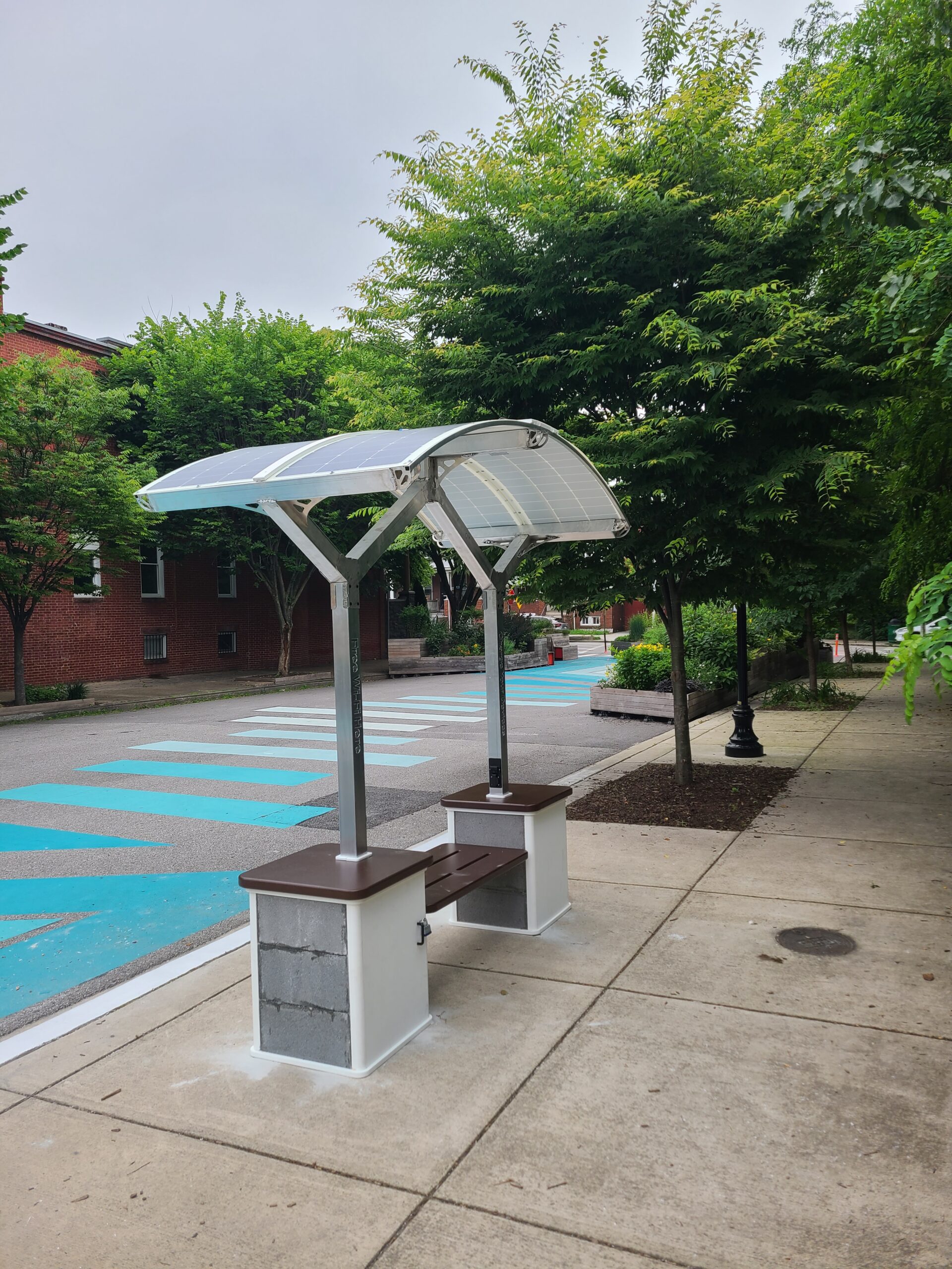 A silver, grey and brown solar-powered WiFi and device charging station by a park with blue lines on grey pavement. 