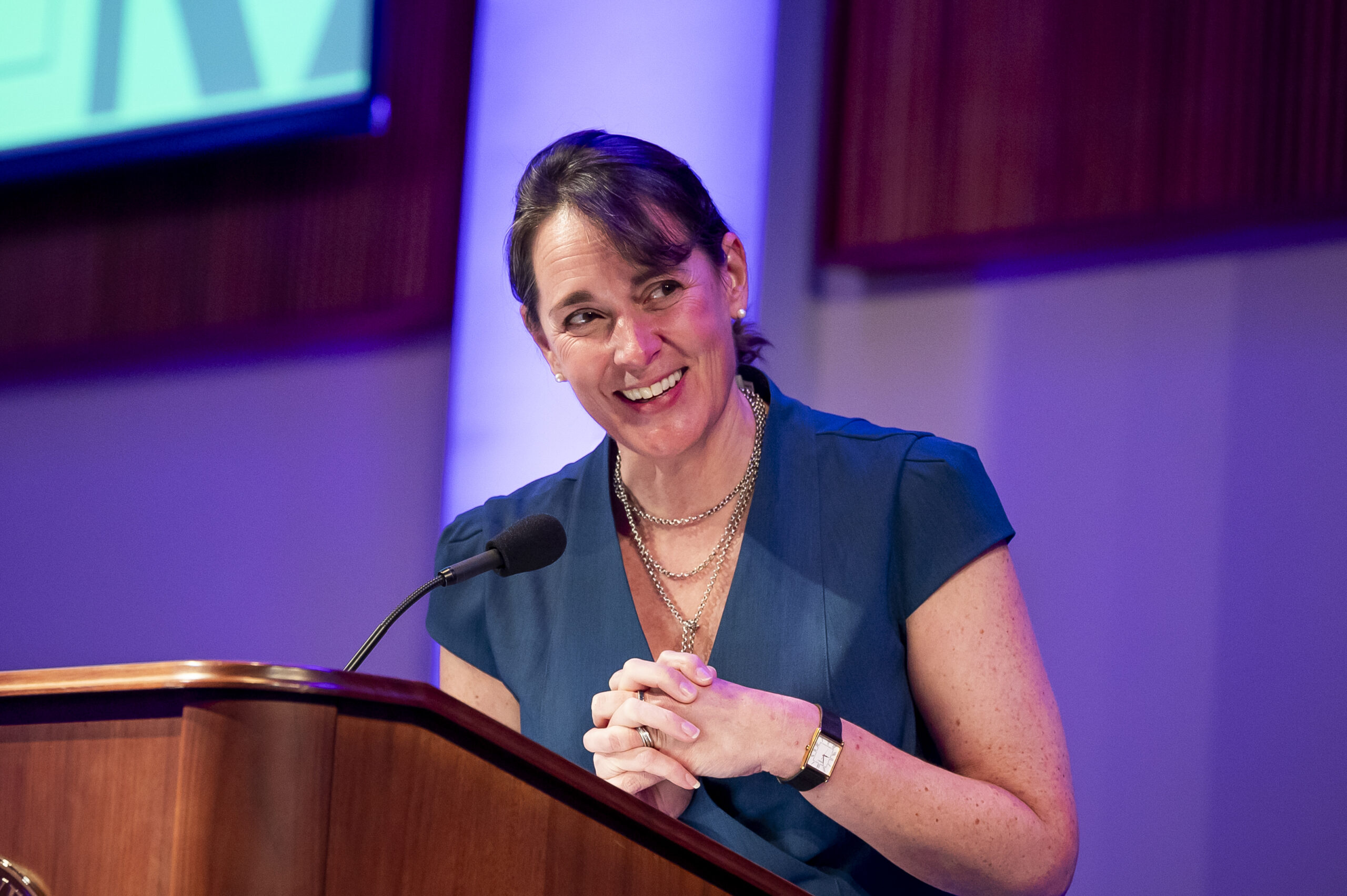 Woman with brown hair smiles at brown podium while wearing teal outfit and silver necklace.