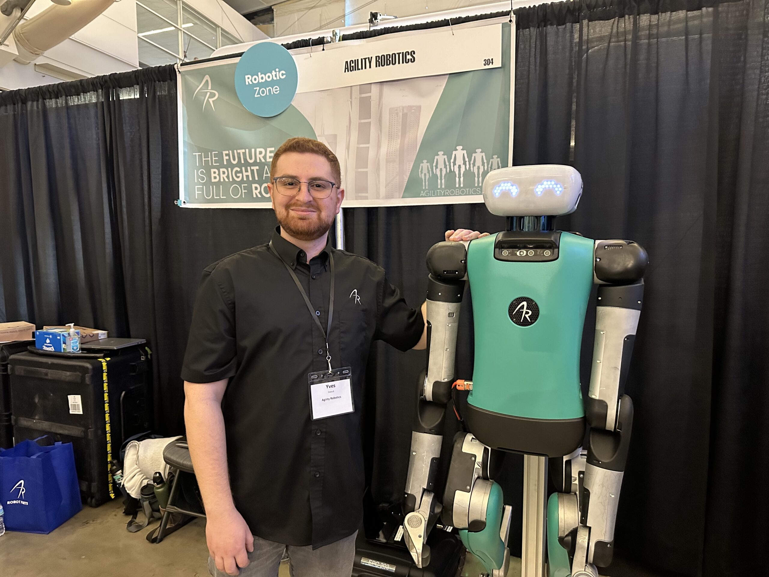 A man stands next to a teal and black robot at a robotics convention. A sign behind them reads "Robotic Zone" and "Ability Robotics.