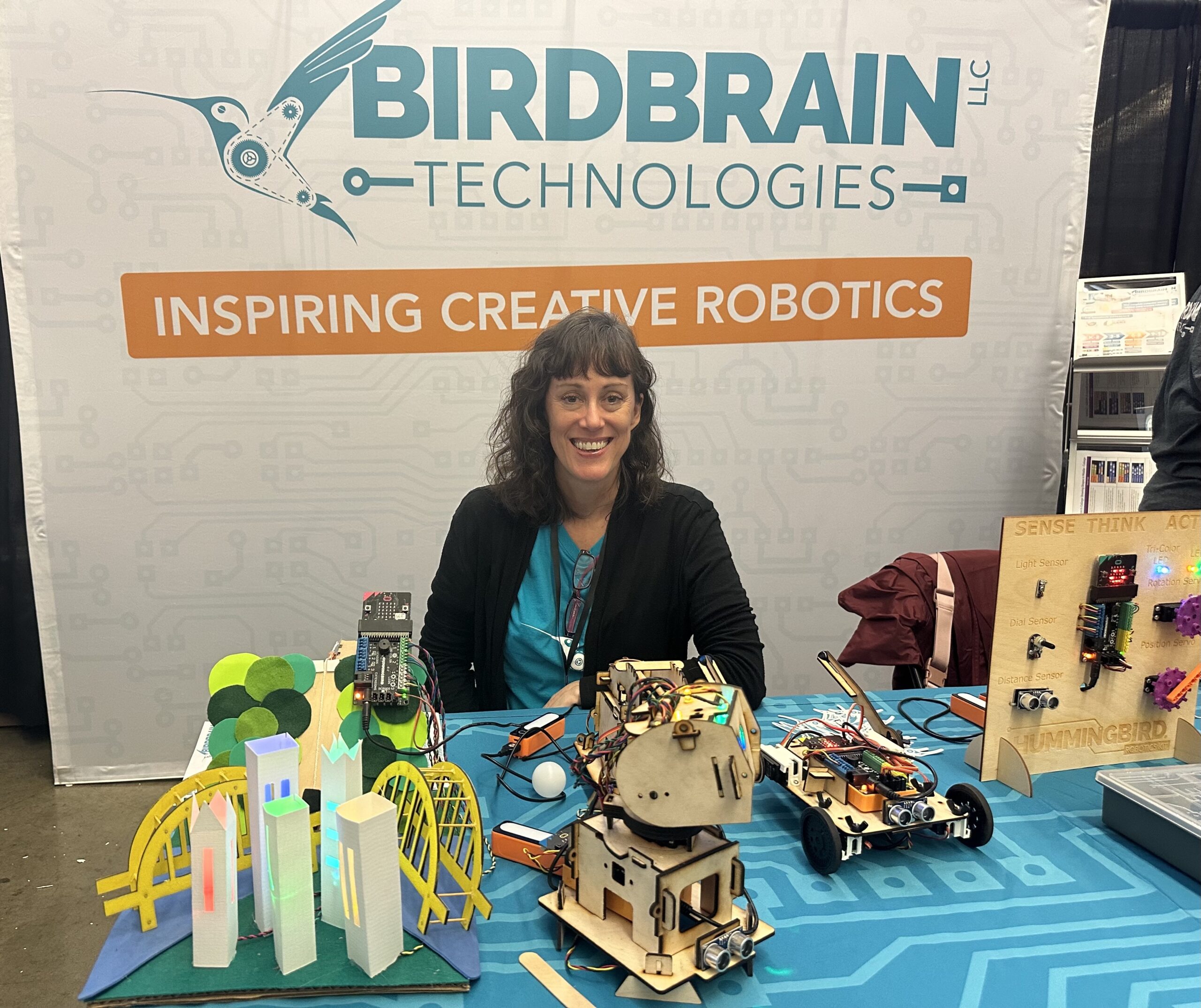 A woman smiles at a booth with robotics displays. The BirdBrain Technologies banner reads "Inspiring Creative Robotics.