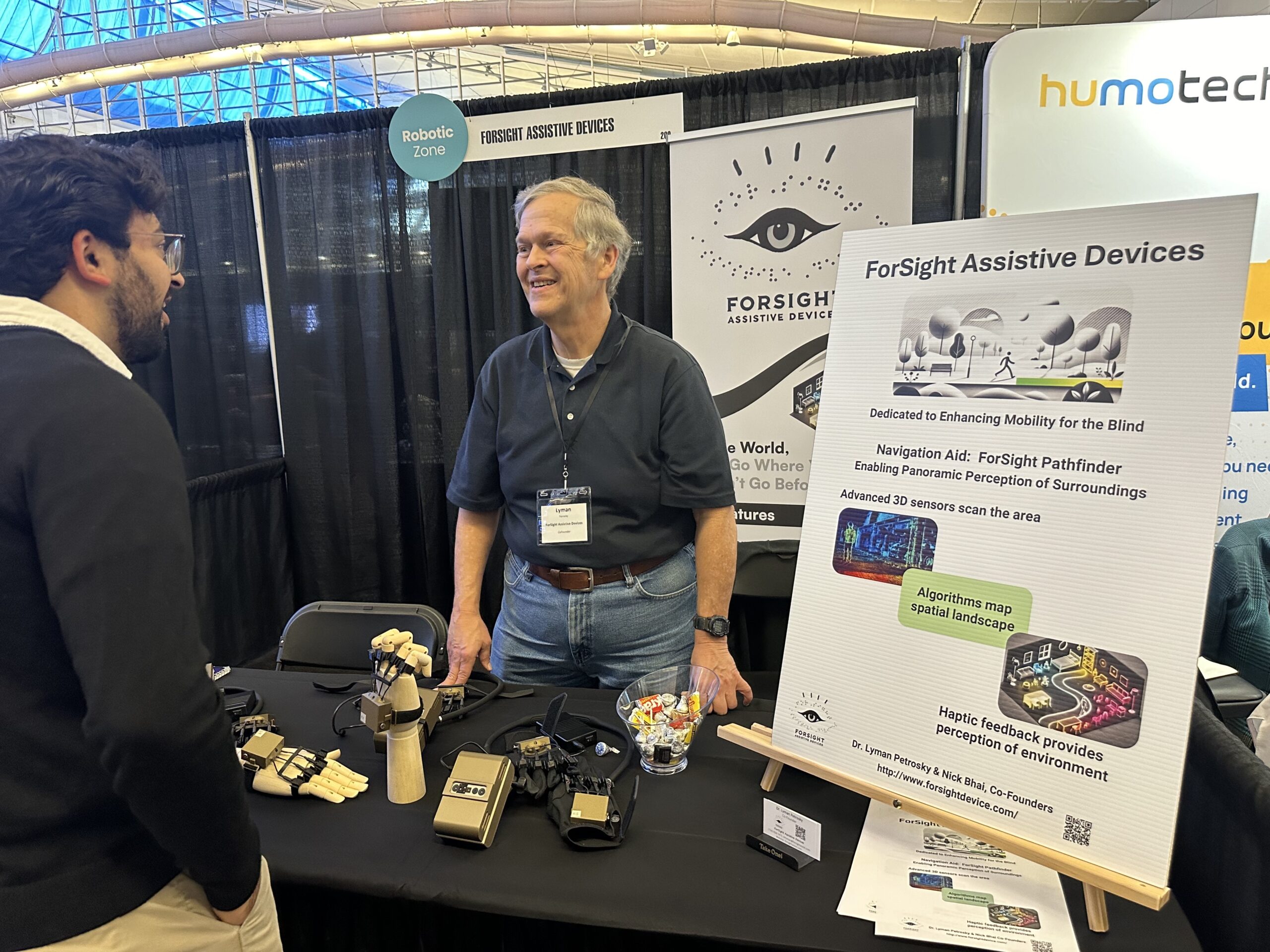 Two men stand near a booth showcasing "ForSight Assistive Devices." A poster describes advanced 3D sensor technology for enhanced mobility for the blind. Devices and candy are on the table.