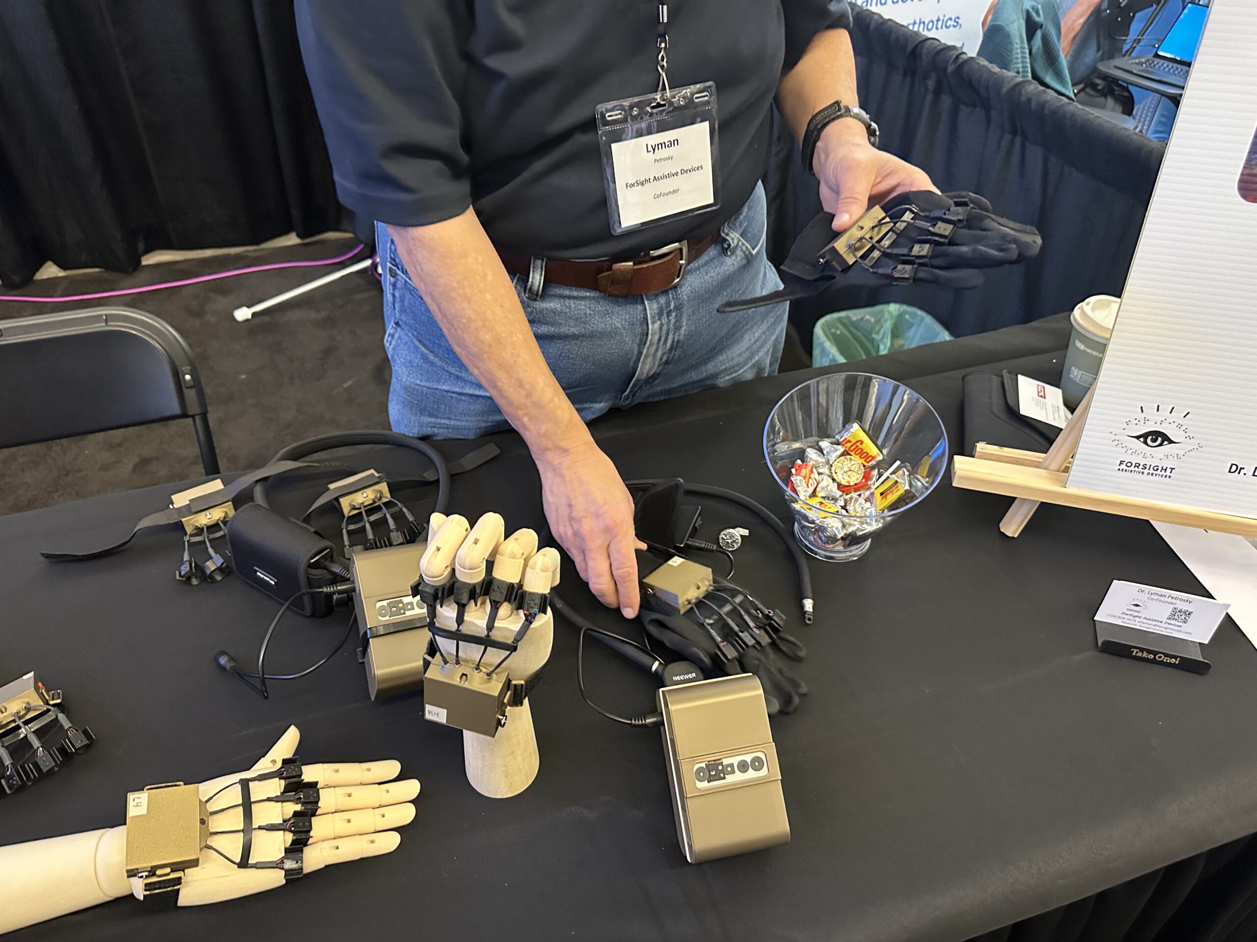 A person demonstrates robotic prosthetic hands on a table, showcasing various models. A bowl of candy and informational materials are also displayed.