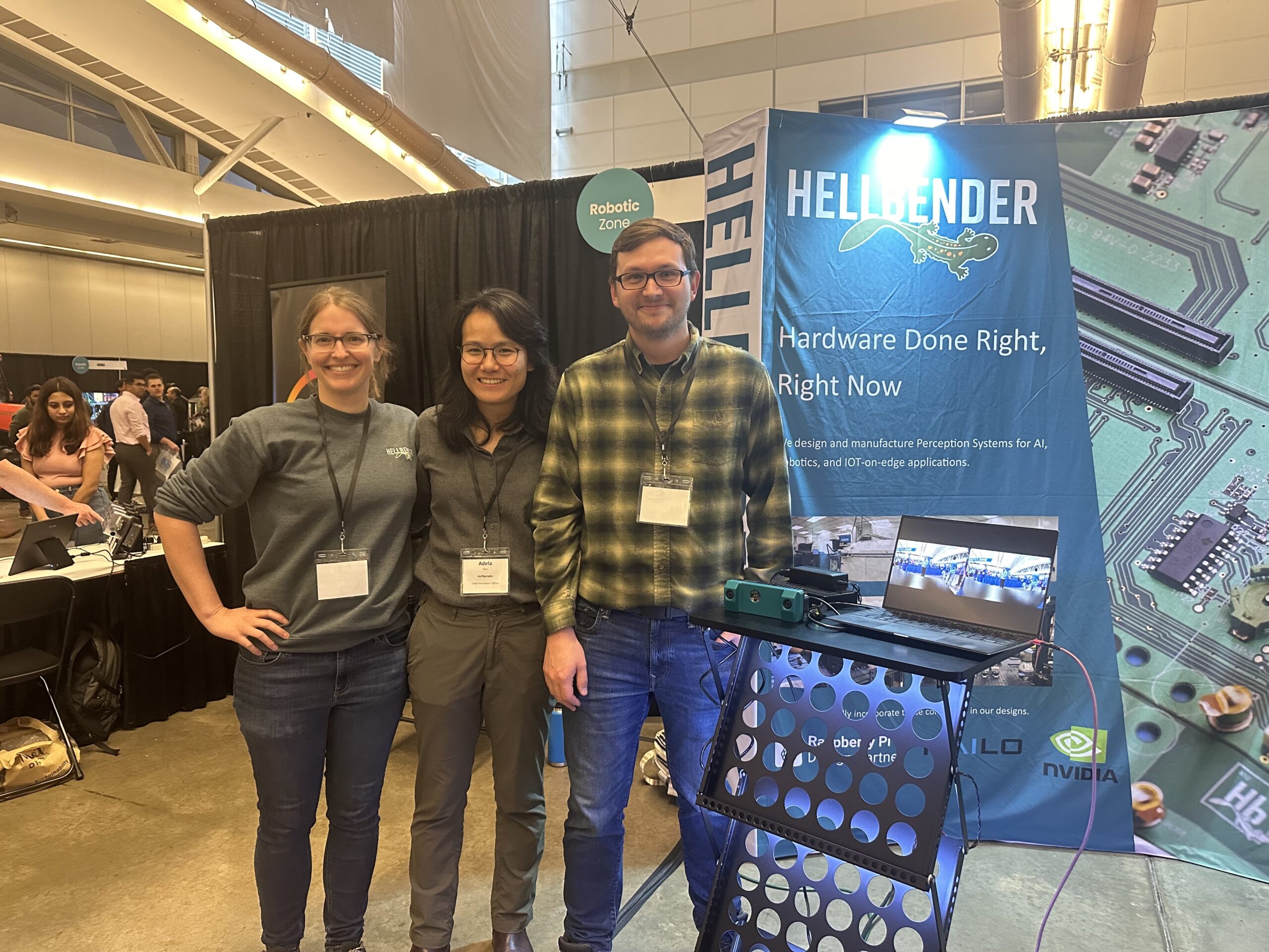 Three people standing in front of a booth with a "Hellbender" banner and tech equipment at a convention.