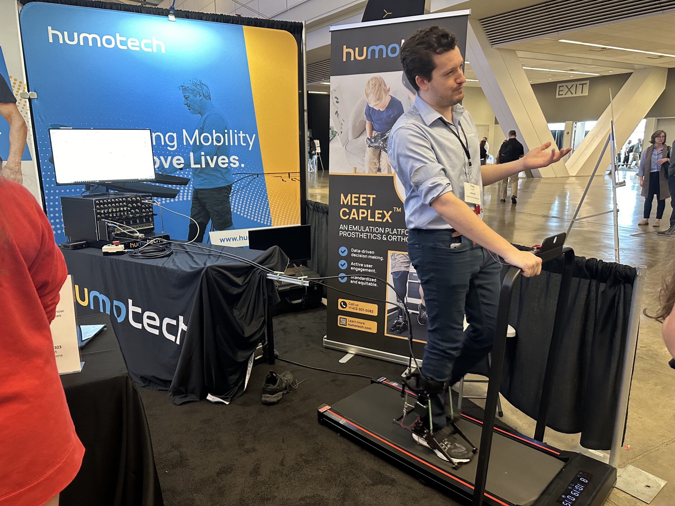 A person demonstrates a mobility device on a treadmill at a Humotech booth during an event. Banners and a computer setup are in the background.