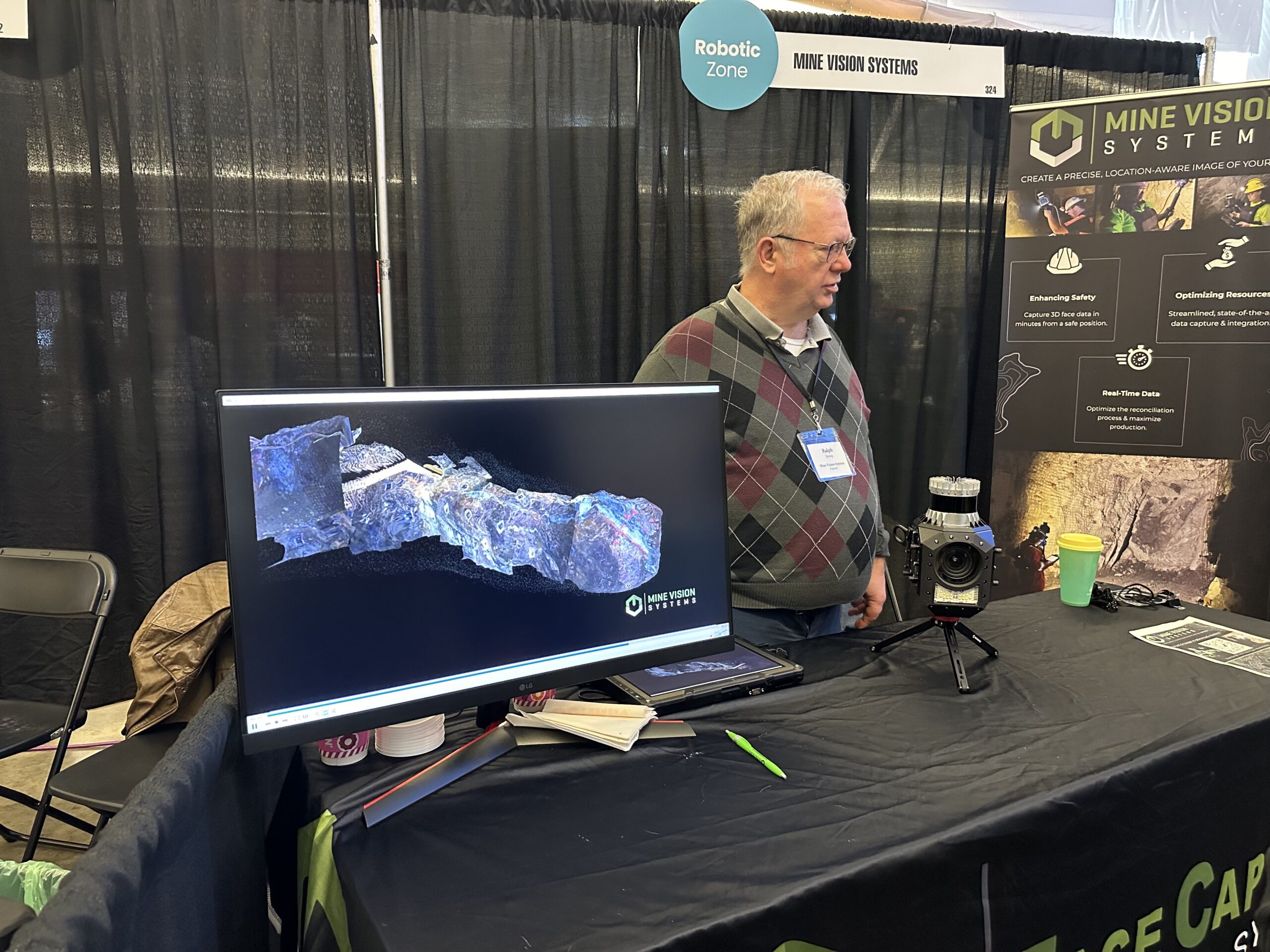 A man stands at an exhibition booth for Mine Vision Systems, featuring a monitor displaying 3D imaging and a camera setup on the table.