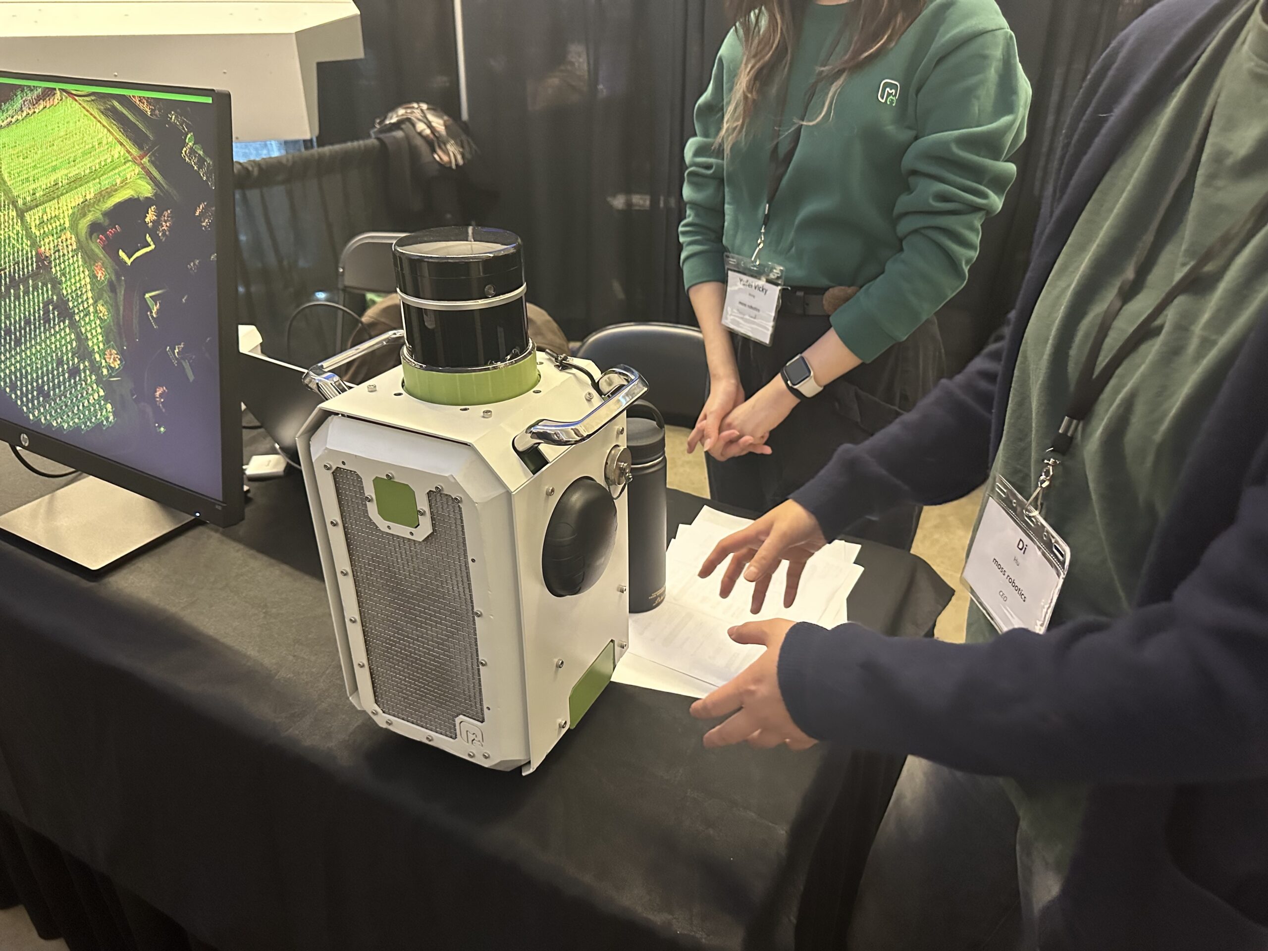 Two people stand by a table displaying a small, box-shaped device with a lens, next to a monitor showing a green aerial image.