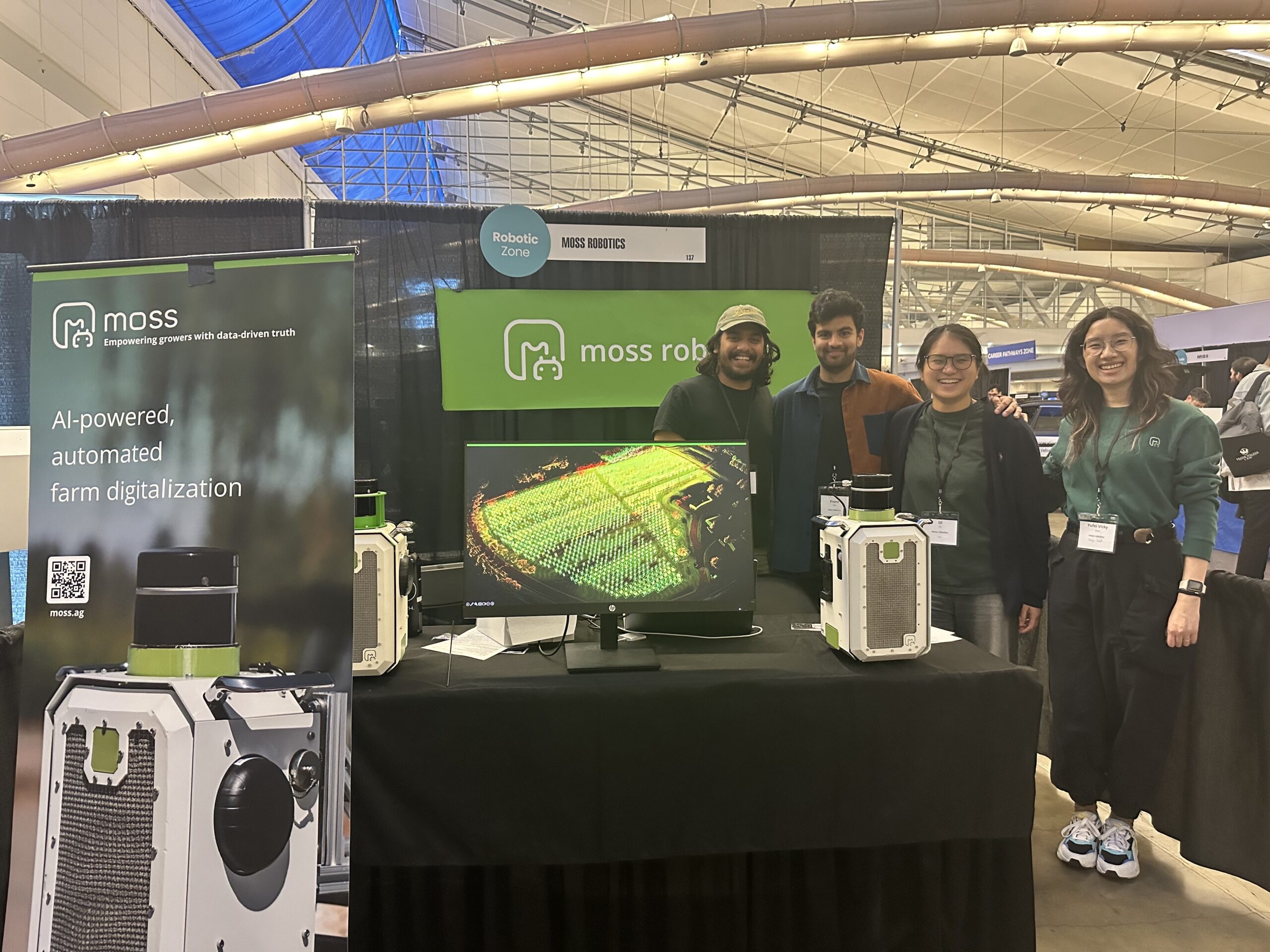 Four people stand at a booth for Moss Robotics, showcasing AI-powered farm digitalization, with a display and equipment on the table.