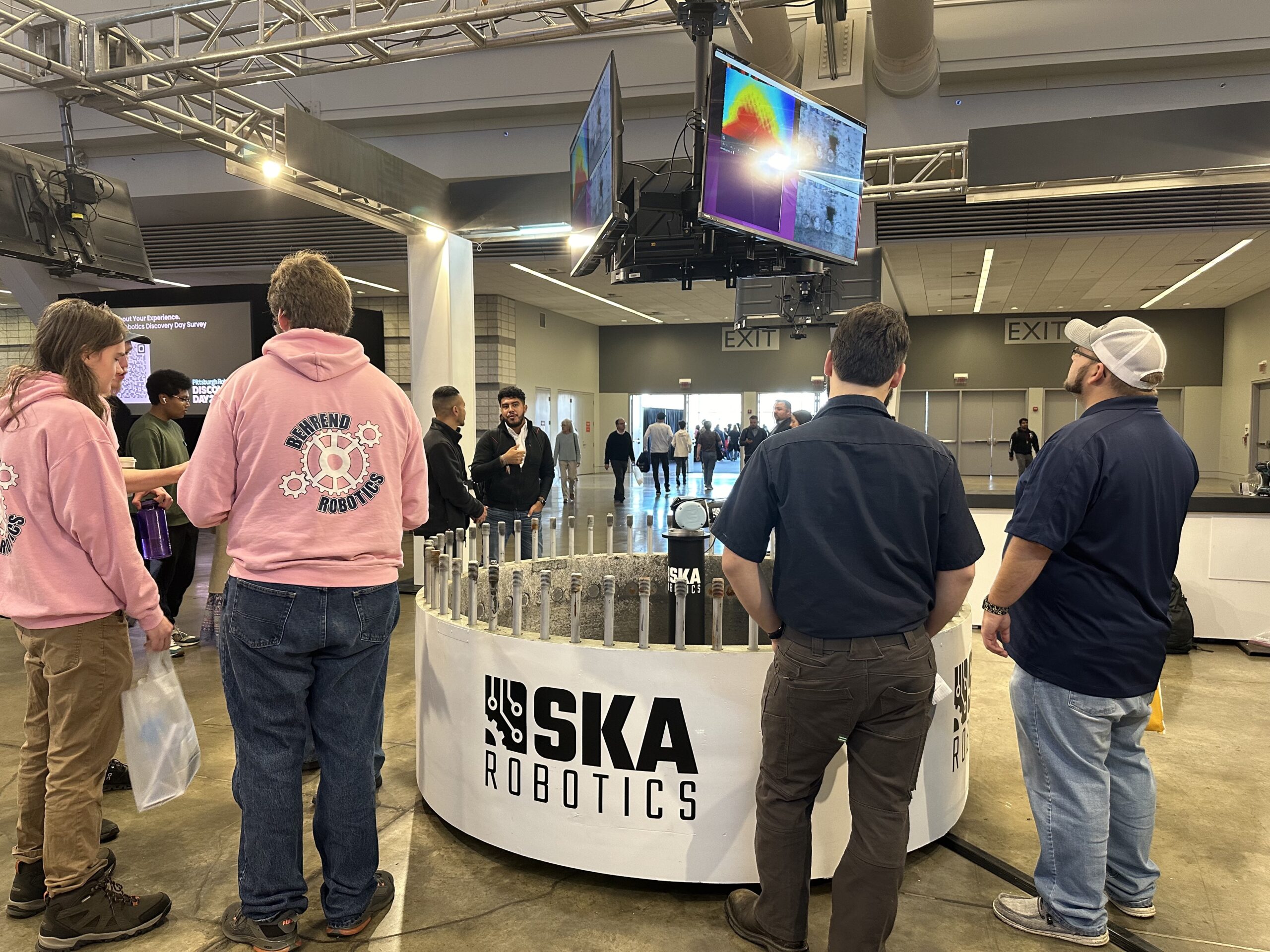 People gather around a display titled "SKA Robotics" at an exhibition. Monitors show a colorful image above.