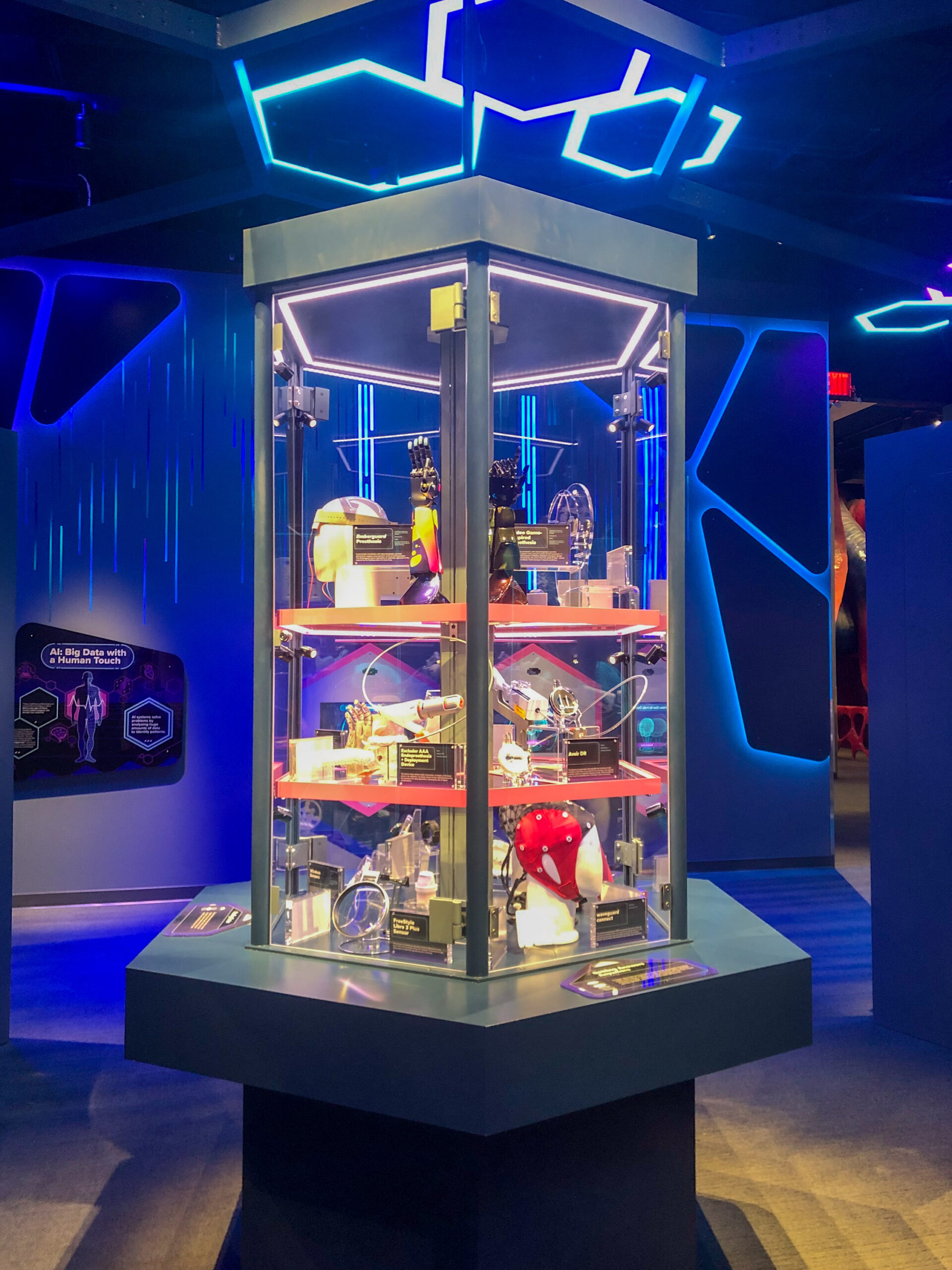 A display case with superhero helmets and props, illuminated by blue and purple lights in an exhibition setting.