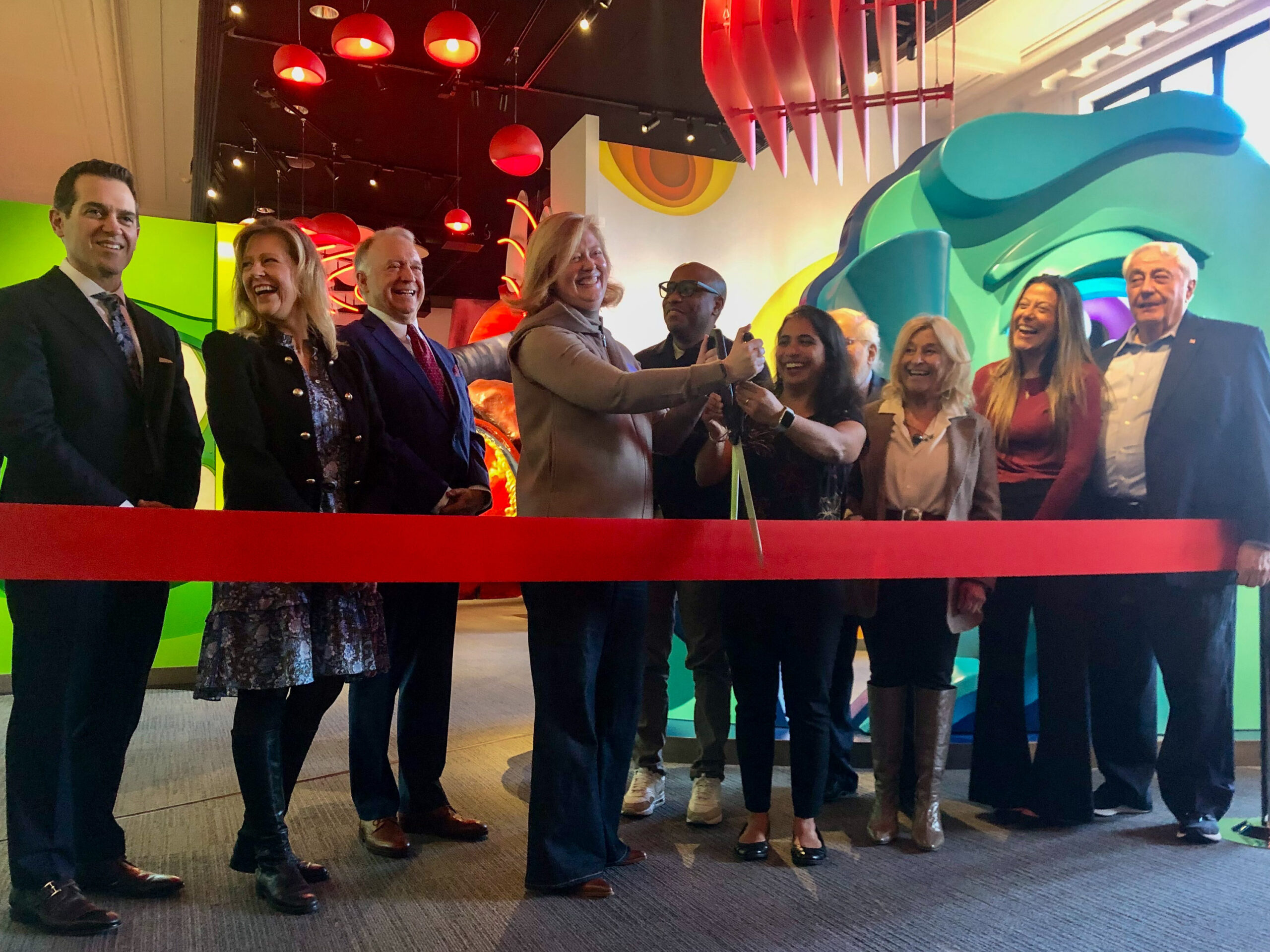 A group of people stands together in a line, cutting a red ribbon at an indoor event with colorful decorations in the background.