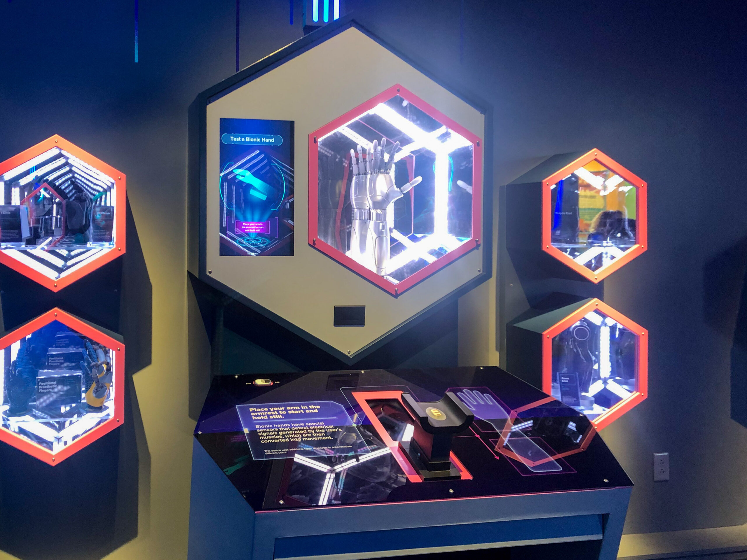 Display of hexagonal cases with futuristic gloves and gadgets, a touchscreen interface, and informational panels in a dimly lit room.