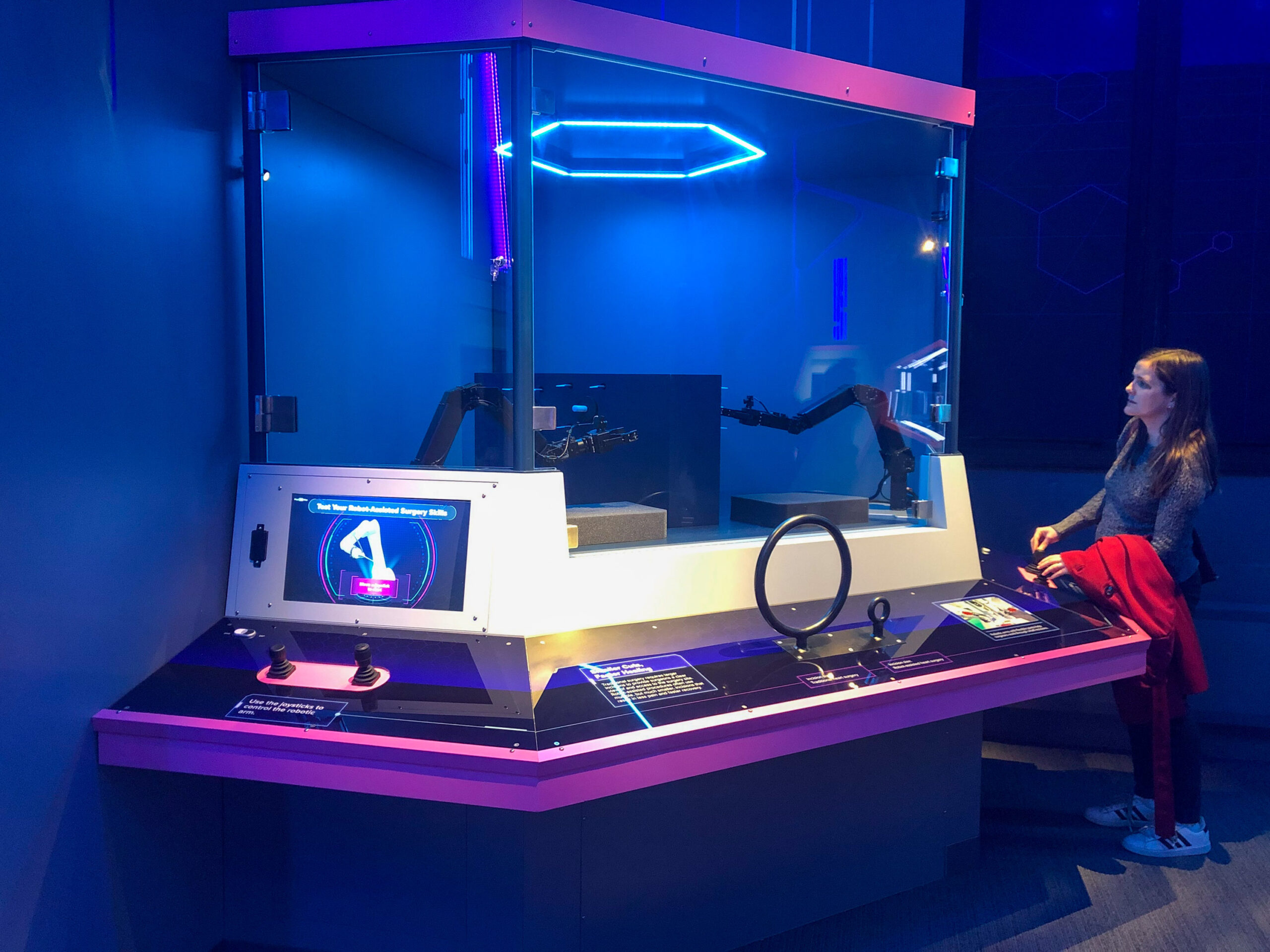 A person observes an interactive touchscreen display and a robotic arm in a futuristic exhibit with blue lighting.