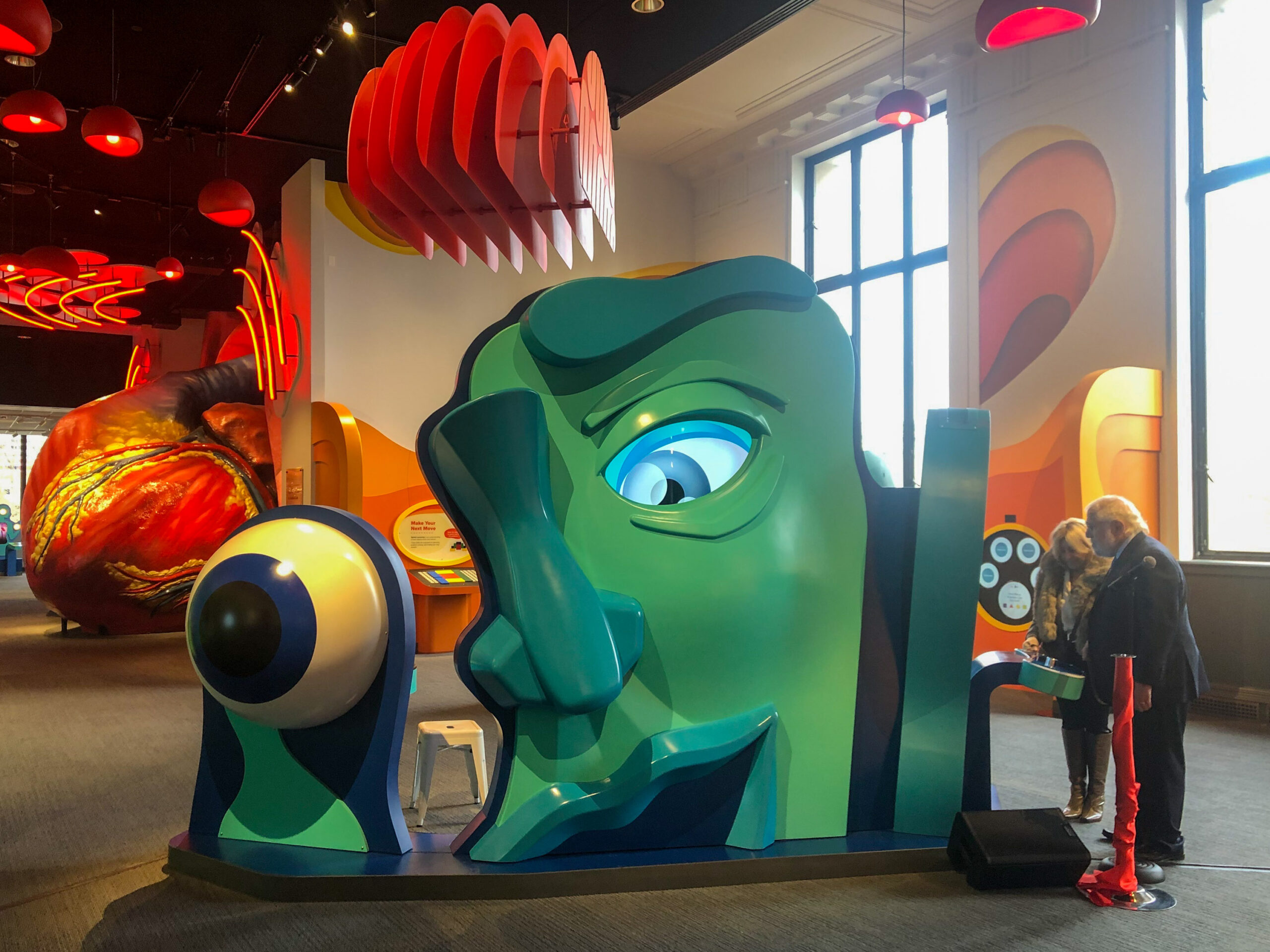 Colorful interactive exhibit with a large green face and eye, designed for visitors to explore. Two people stand nearby in a brightly lit room with abstract decorations.