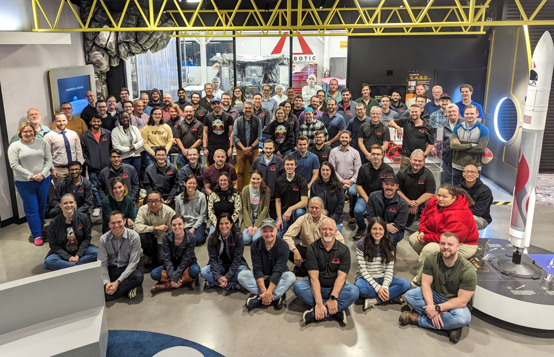 A large group of people stand or sit in front of a clear glass window. Behind the window is Astrobotic's Peregrine Mission One. 