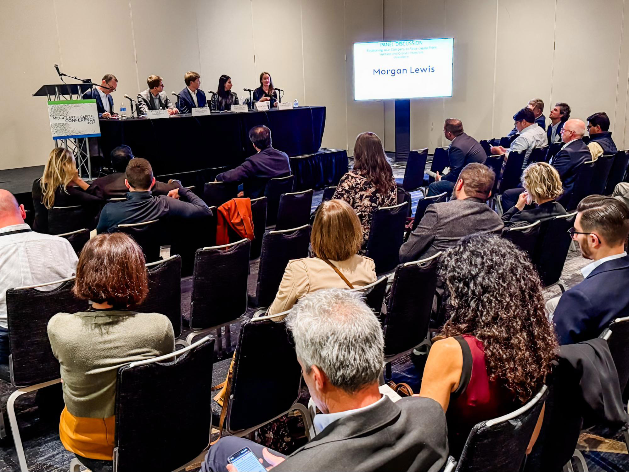 Audience attending a panel discussion in a conference room. A sign reads "Morgan Lewis" on a screen behind the panelists.