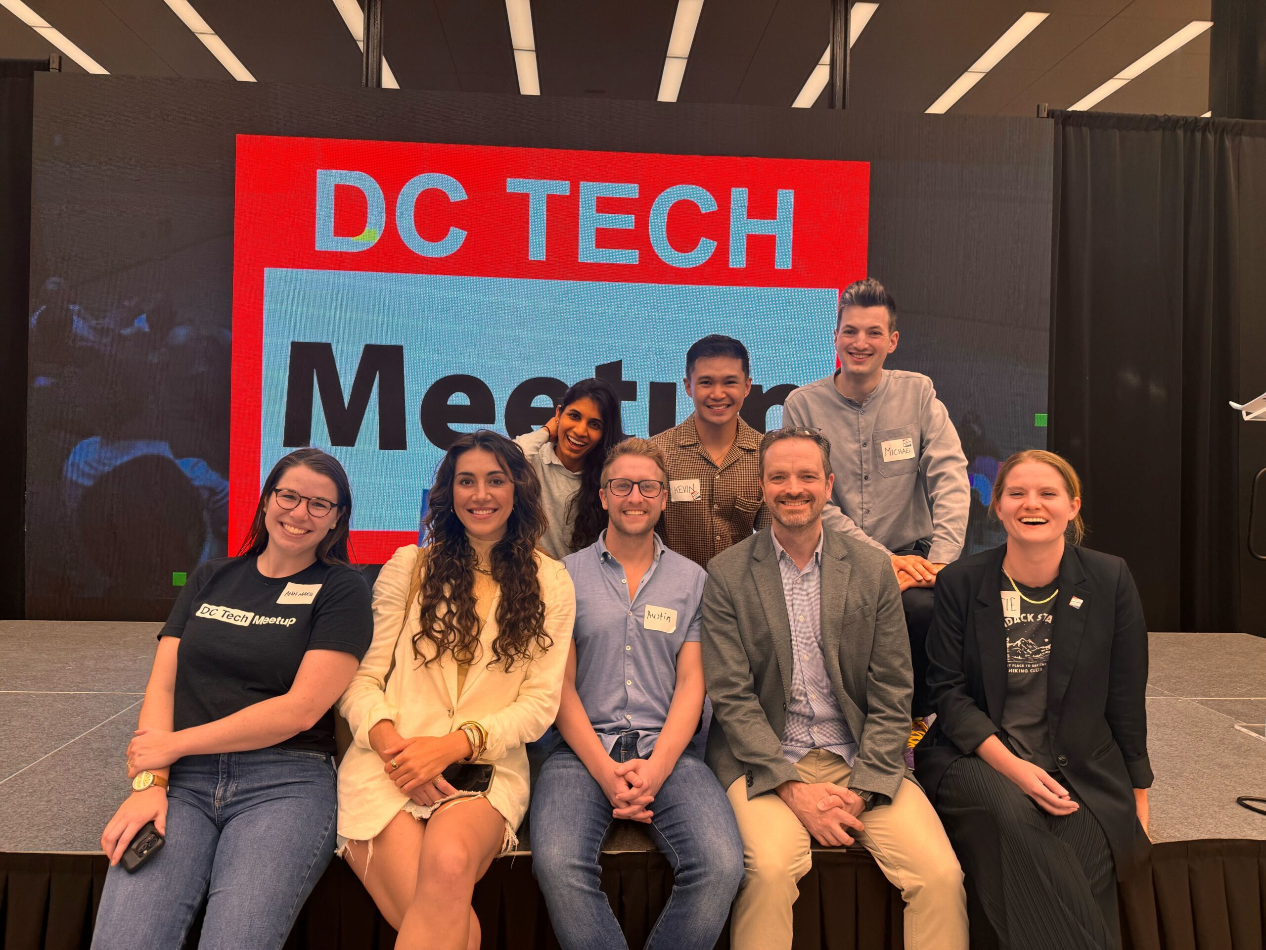 Eight people are posing together on stage in front of a screen displaying the text "DC Tech Meetup."