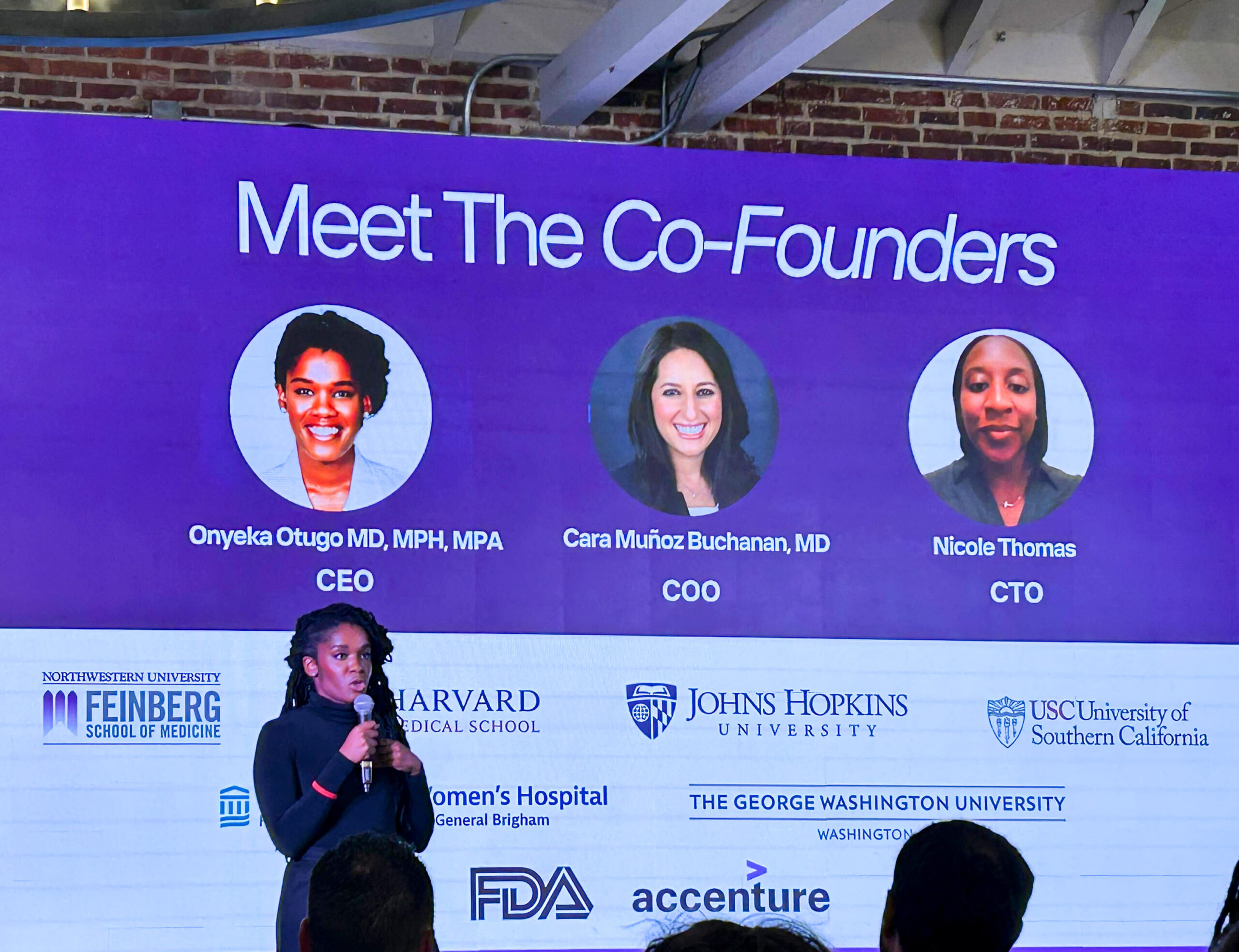 A Black woman presents to audience before screen with presentation on purple-lit red brick wall.