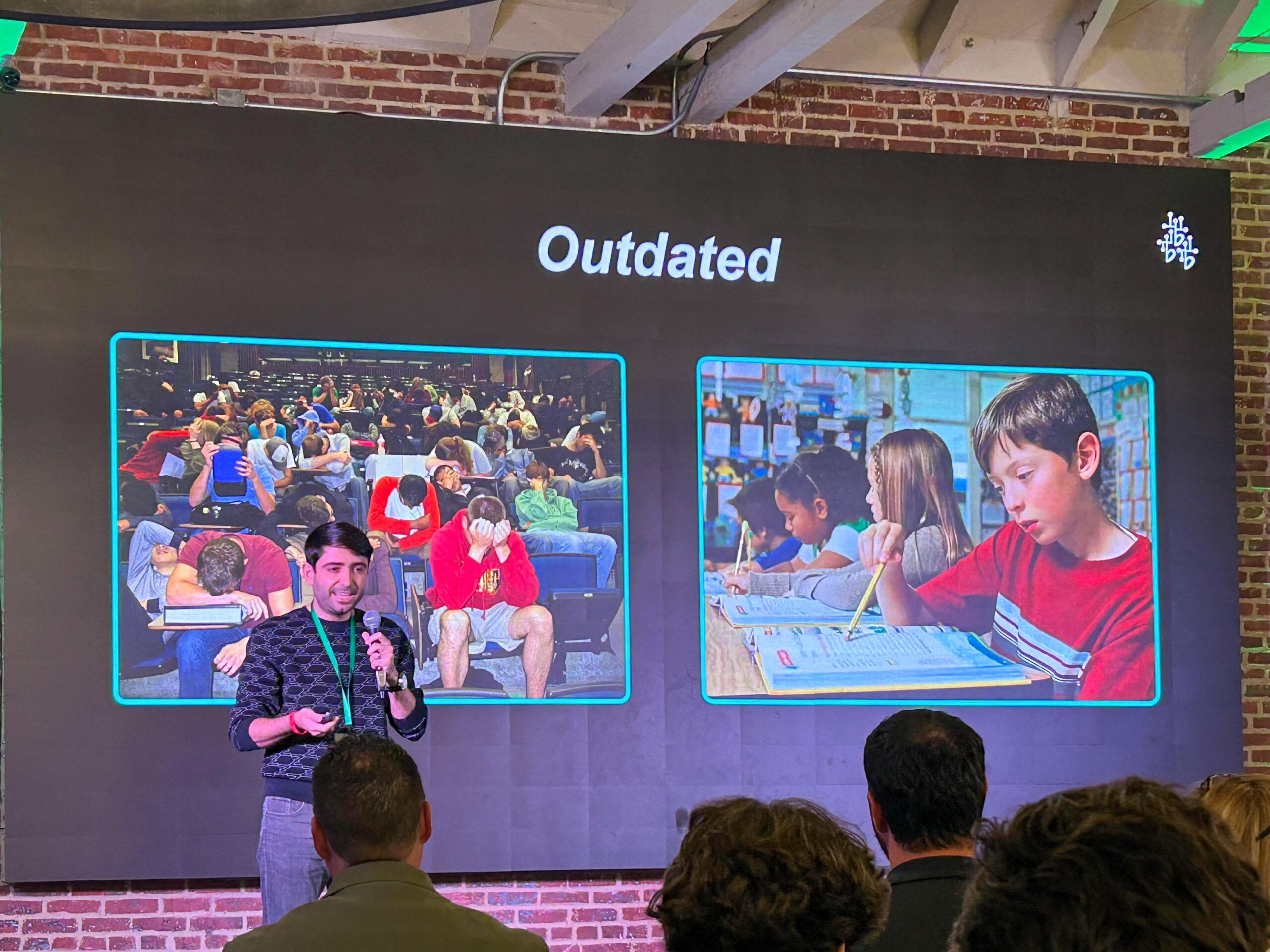 A man presents to audience before screen with presentation on purple-lit red brick wall.