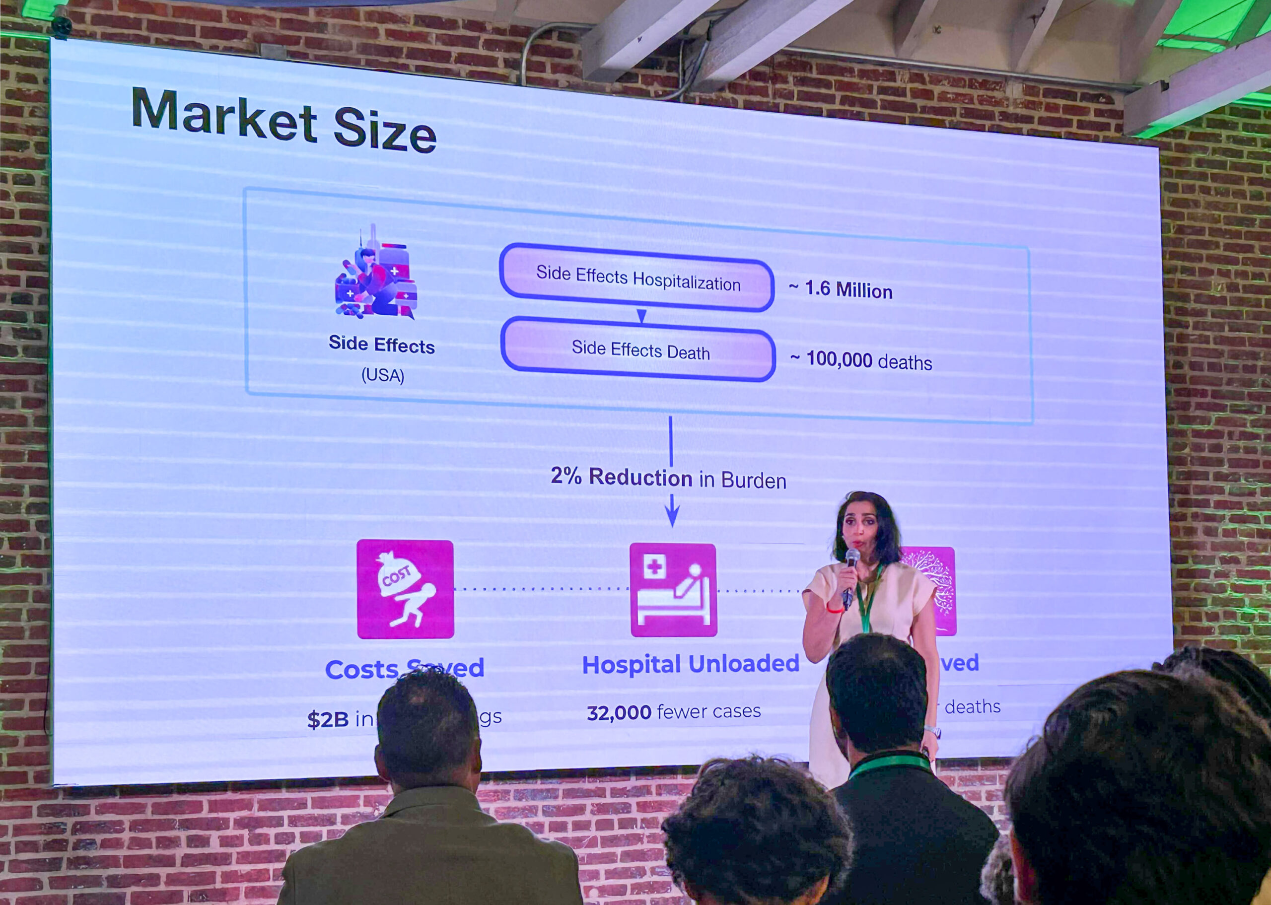 A MENA woman presents to audience before screen with presentation on purple-lit red brick wall.