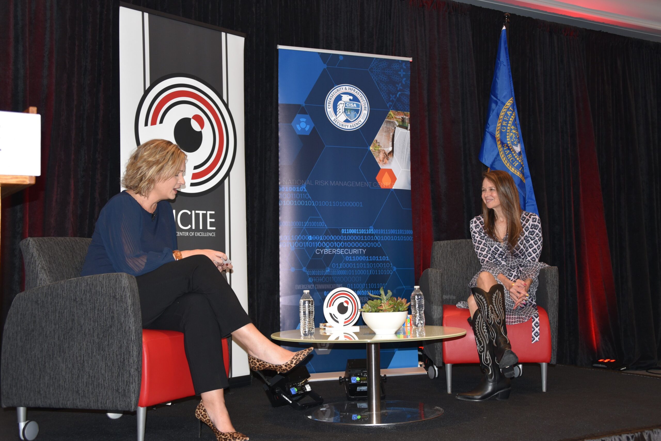 Two white women sit on red chairs by brown coffee table and blue and black curtains and signage. 